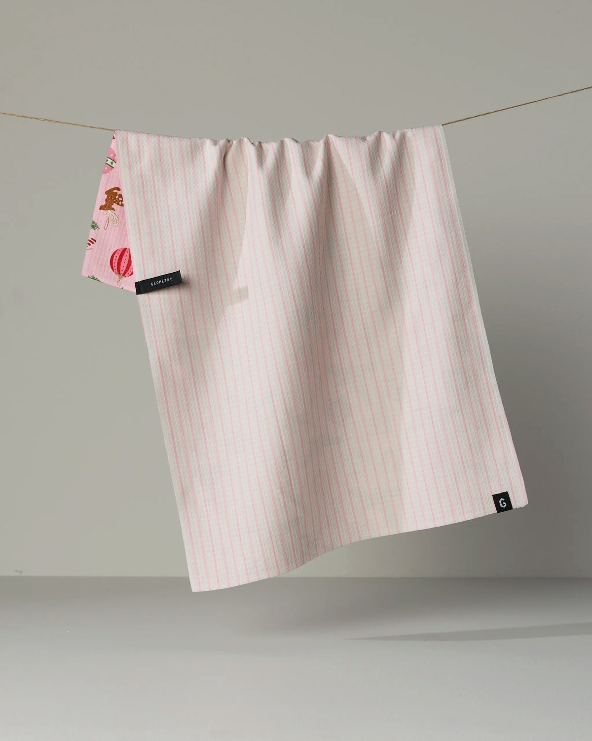 The Pink Tidings tea towel by Geometry, crafted from recycled materials, features light pink and white stripes and a small pink macaron detail in one corner. It hangs from a string against a plain gray background.