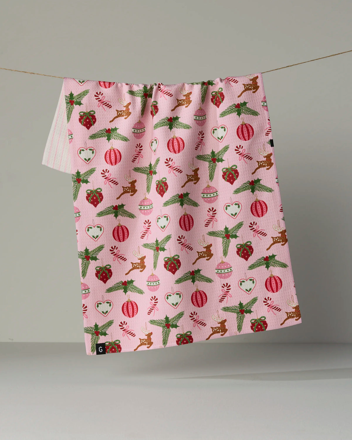 Introducing Pink Tidings by Geometry—a double-sided tea towel crafted from recycled materials. Showcasing a festive pink design adorned with Christmas-themed patterns such as reindeer, hearts, holly, candy canes, and ornaments, this quick-drying tea towel blends eco-friendly functionality with holiday charm. Shown here hanging on a clothesline against a neutral background.