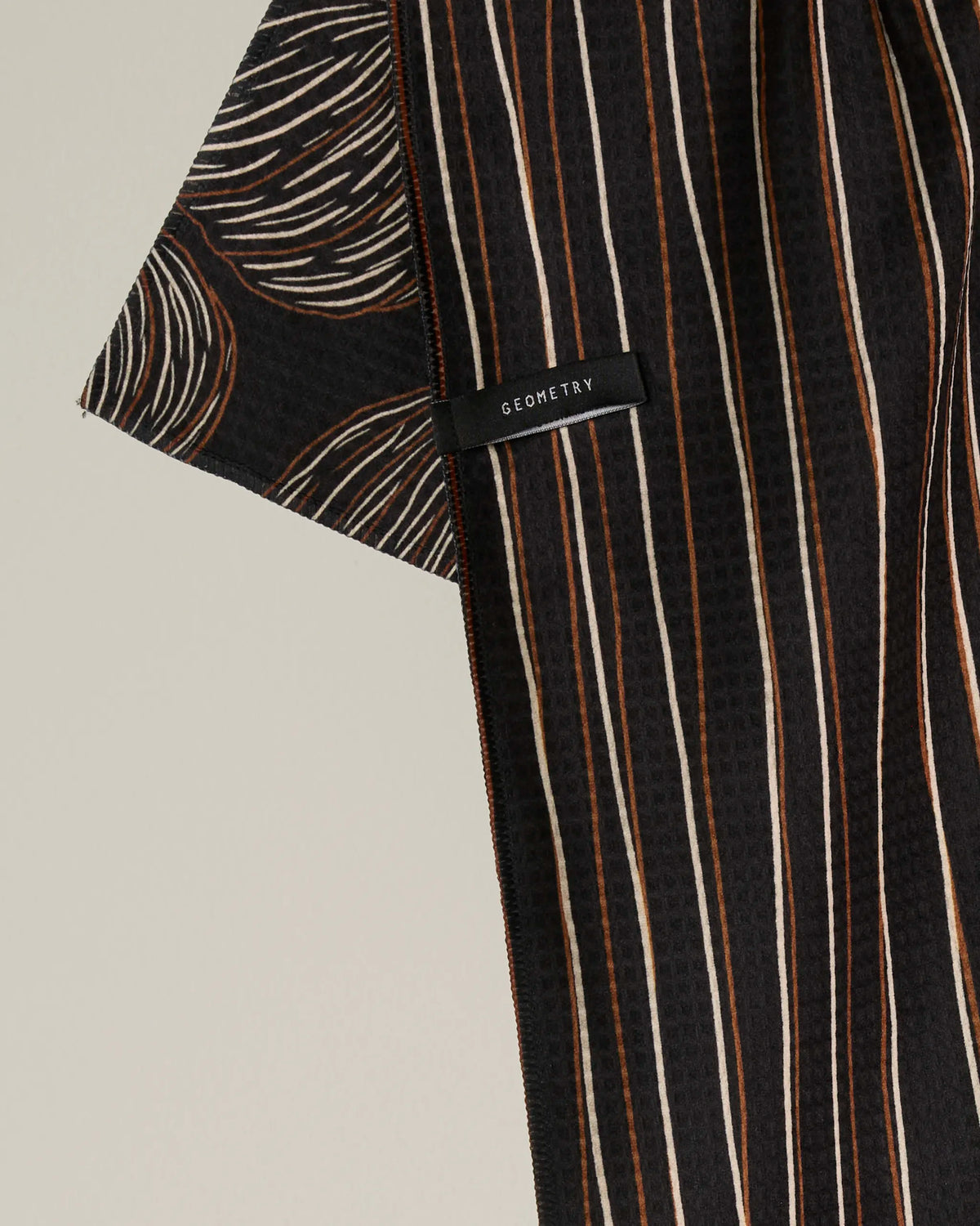 A close-up of the “Pinstripe Patch” recycled kitchen towel by Geometry, featuring vertical beige and brown stripes with brown curved lines on a black background. A small black tag with “GEOMETRY” in white text is sewn onto the fabric.