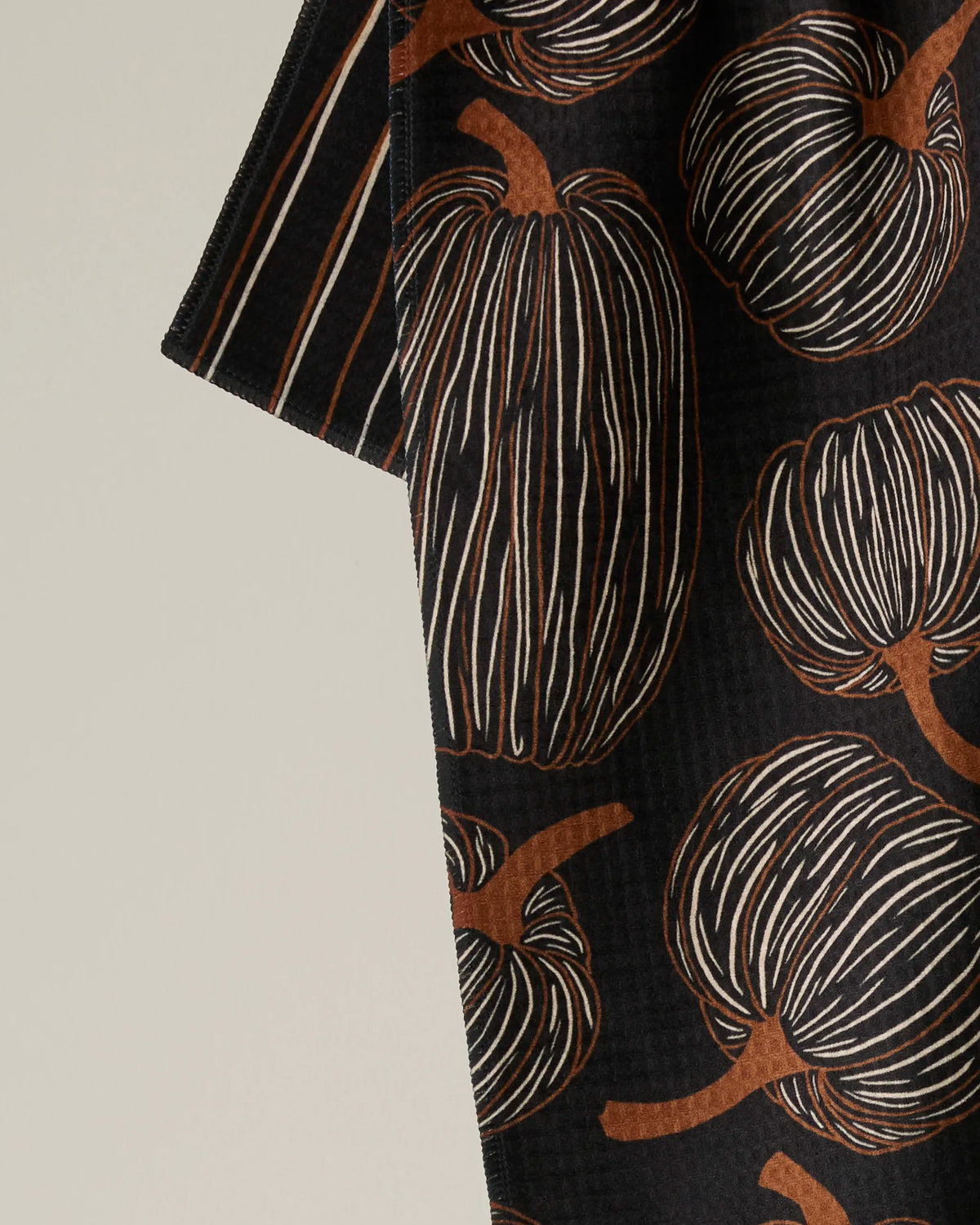 Introducing Pinstripe Patch by Geometry—a double-sided tea towel adorned with a distinctive pattern of brown and white striped pumpkins on a sleek black fabric, artfully draped against a light background. This stylish tea towel infuses your home with autumnal charm and versatile functionality.