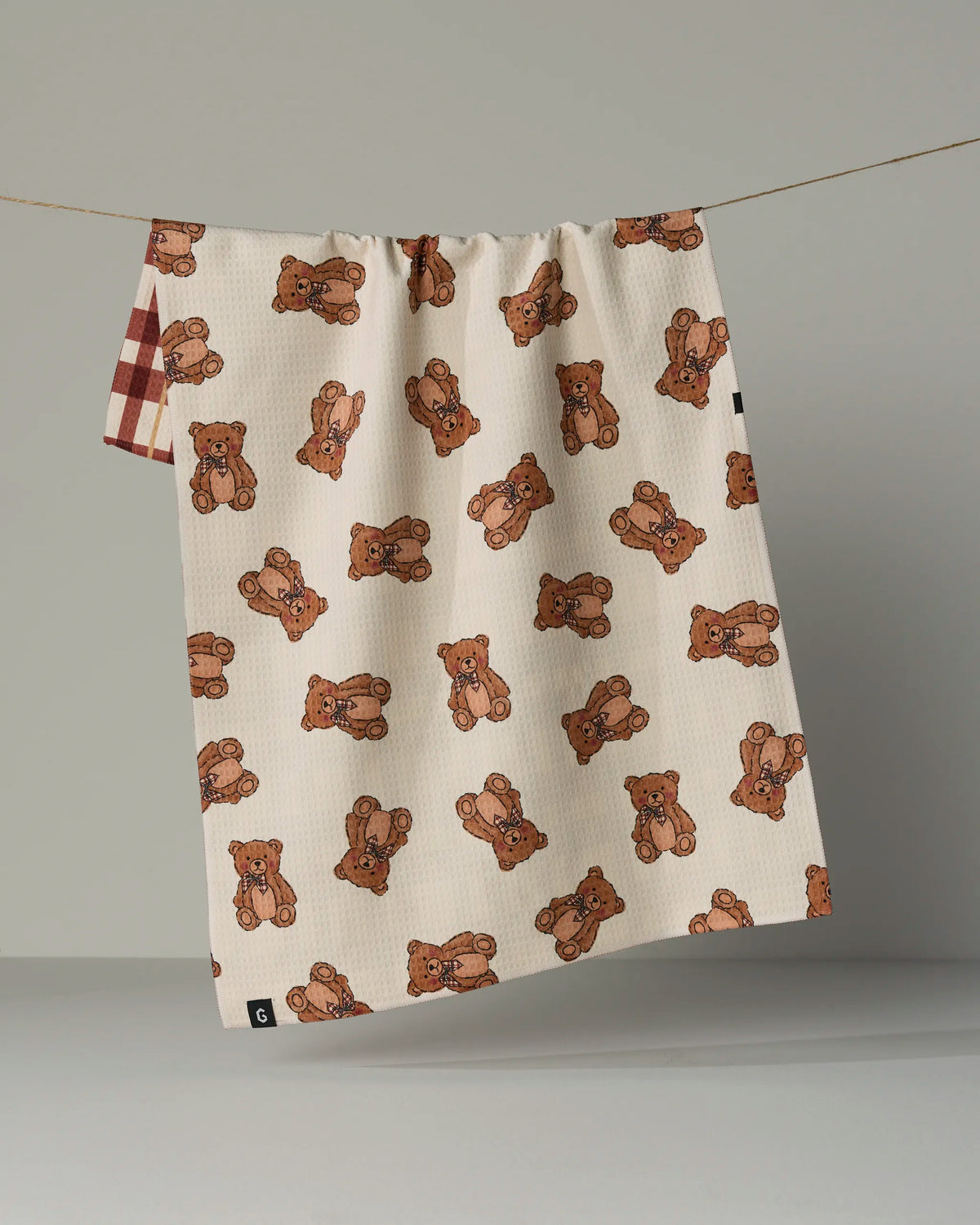 The Plaid Teddy tea towel by Geometry, crafted from recycled materials, features a double-sided design: one side adorned with an adorable brown teddy bear pattern and the other corner accented with a red and white plaid. Absorbent and quick-drying, this standout towel hangs on a clothesline against a simple neutral background.
