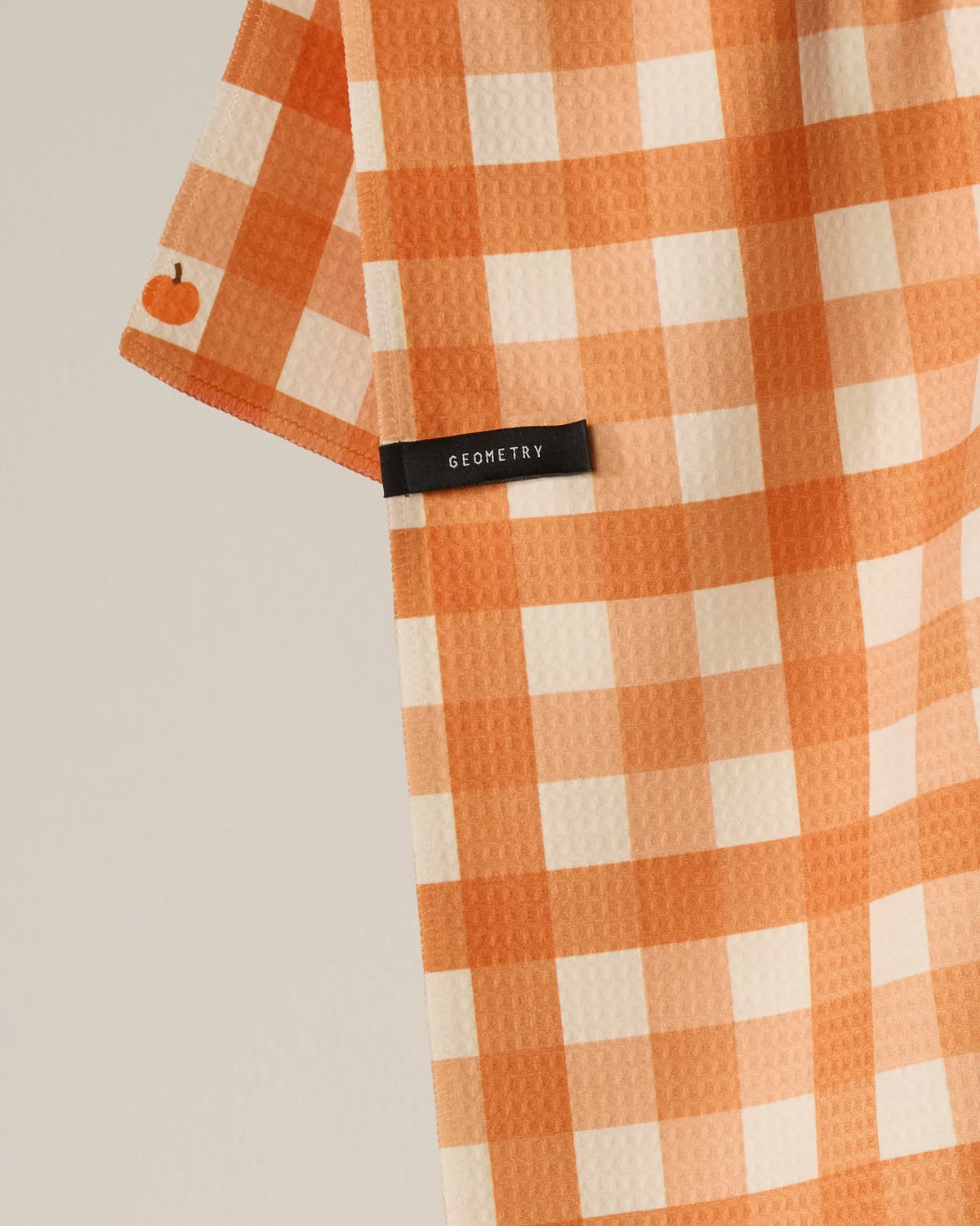 A recycled kitchen towel from Geometry, called Pumpkin Check, hangs against a light background. It features an orange and white checkered pattern, a small pumpkin icon on the edge, and a black label with the brand name Geometry.