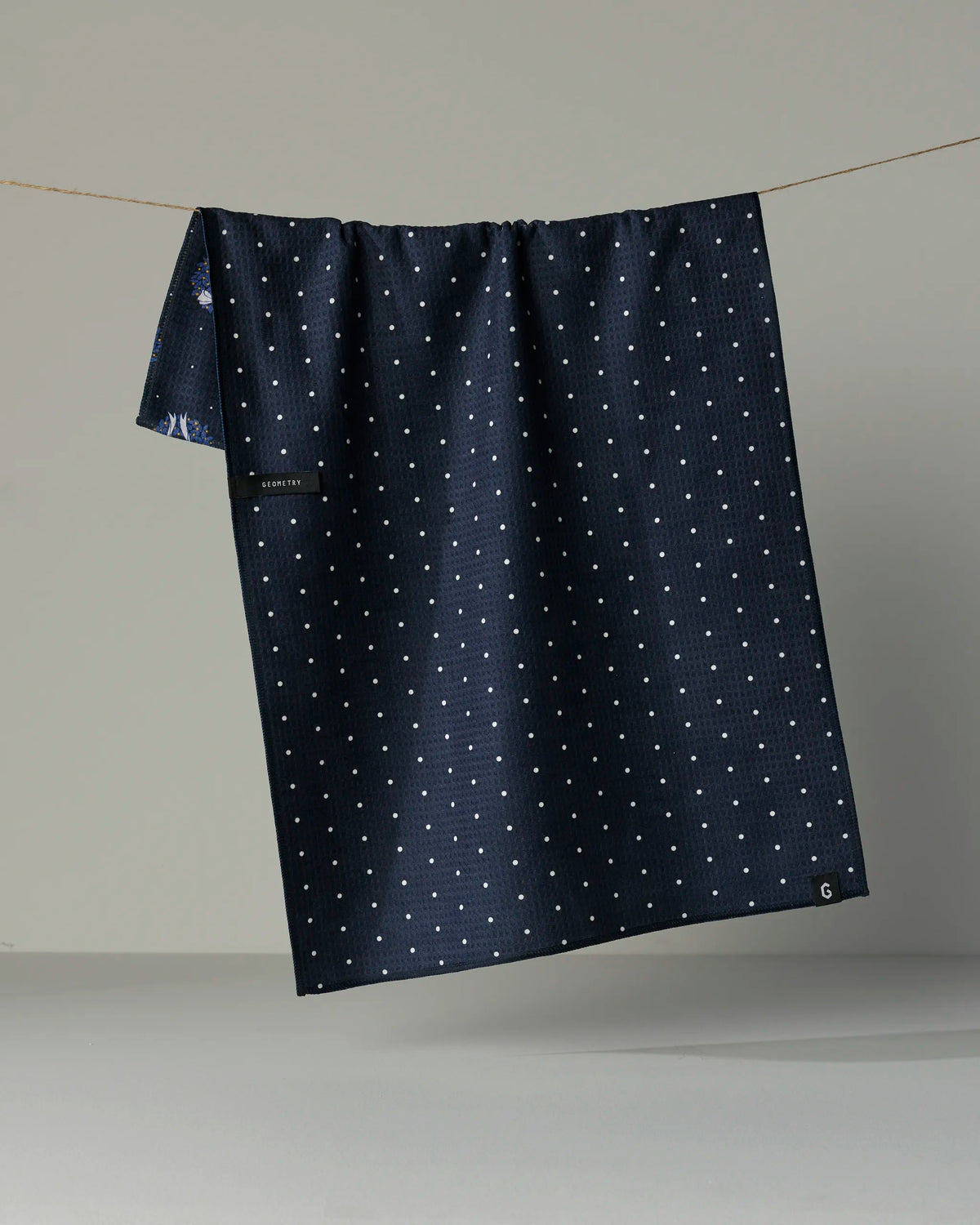 The Quiet Skies tea towel by Geometry, featuring a dark blue double-sided design with a small white square pattern, hangs from a thin string against a plain gray background.