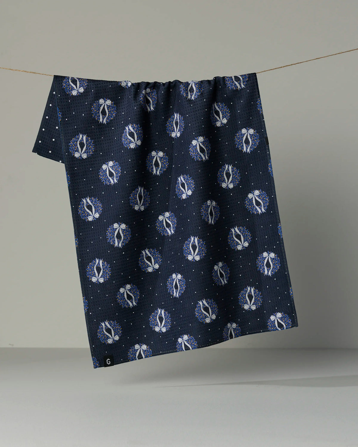 A Quiet Skies tea towel by Geometry, featuring a double-sided dark blue design with a repeating white astronaut pattern, hangs on a string against a plain gray background.