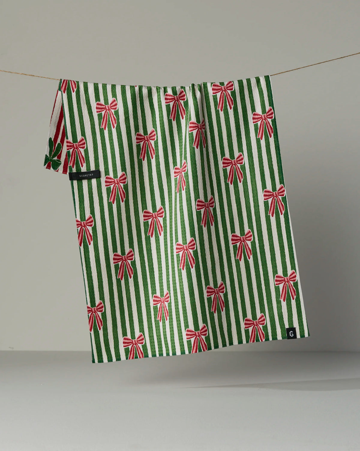 A double-sided tea towel from Geometry, called “Ribbon Bows,” featuring green and white stripes with a pattern of repeating red and white ribbon bows, hangs on a line against a neutral background.
