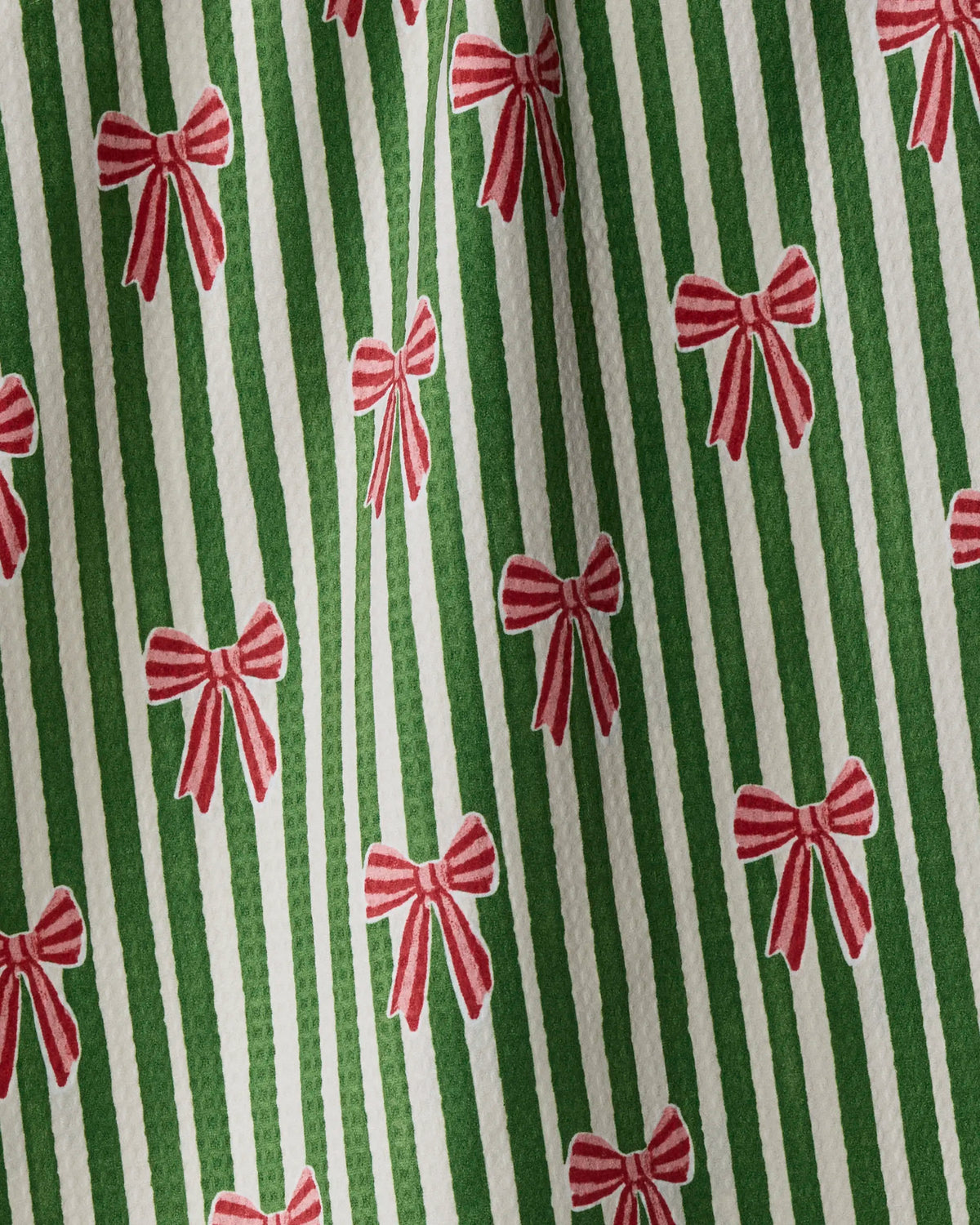 The “Ribbon Bows” tea towel by Geometry features green and white vertical stripes adorned with small red and white bows. The double-sided design appears slightly wrinkled, casting gentle shadows and adding texture to the image.