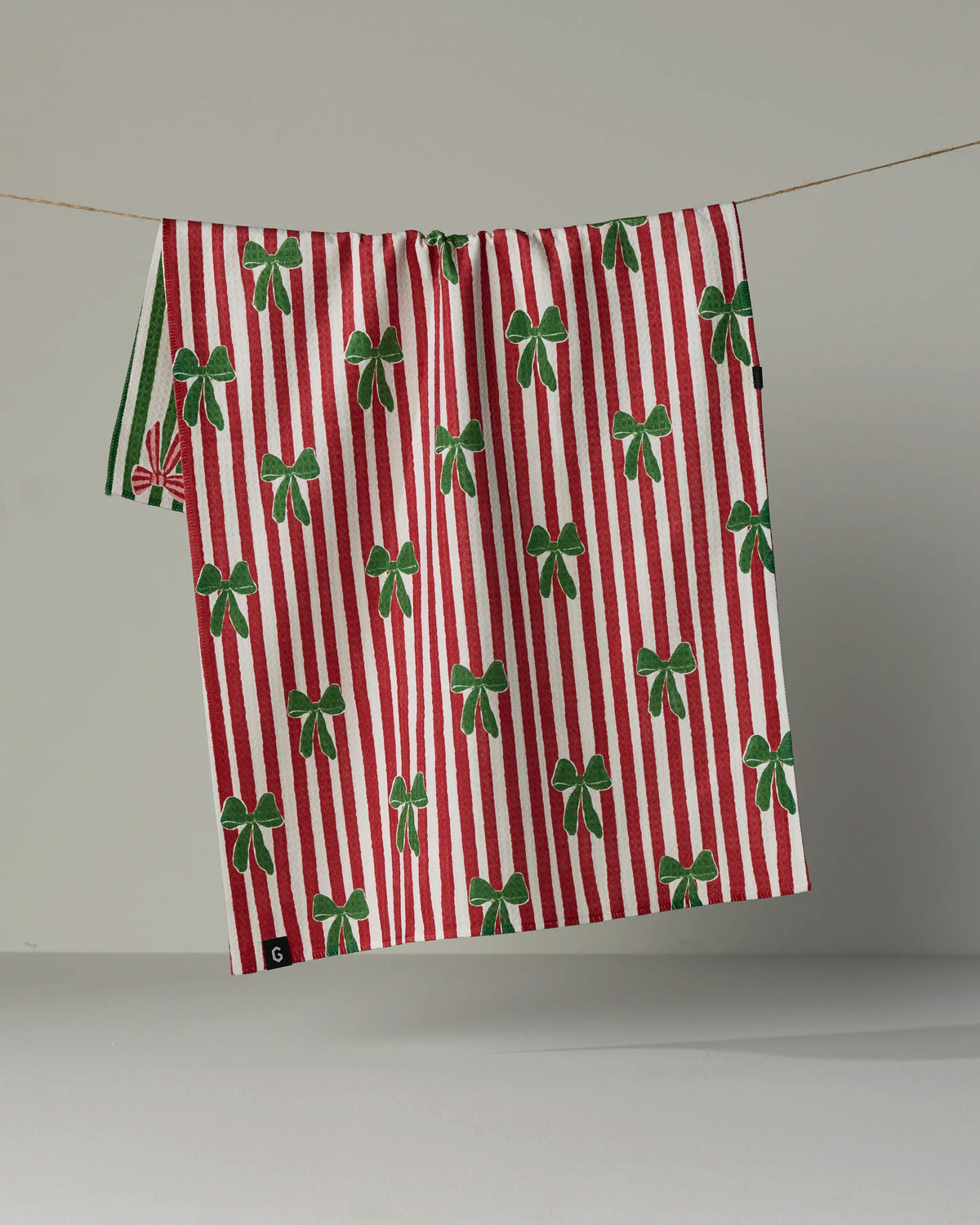 A double-sided tea towel from Geometry’s Ribbon Bows collection, featuring red and white vertical stripes with green bow patterns, hangs on a clothesline against a plain gray background.