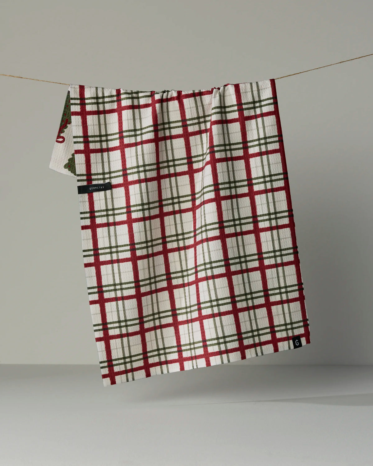 A “Ribbon Pines” towel by Geometry, featuring a red and green plaid pattern, hangs on a string against a plain light gray background.