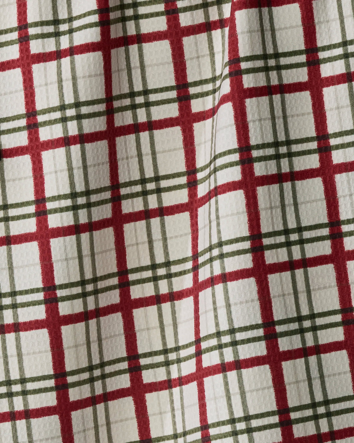 A close-up of the Ribbon Pines tea towel by Geometry showcases its red and green plaid pattern on a white background. The textured fabric and gently wavy lines highlight the folds in this double-sided towel, which is made from recycled materials.