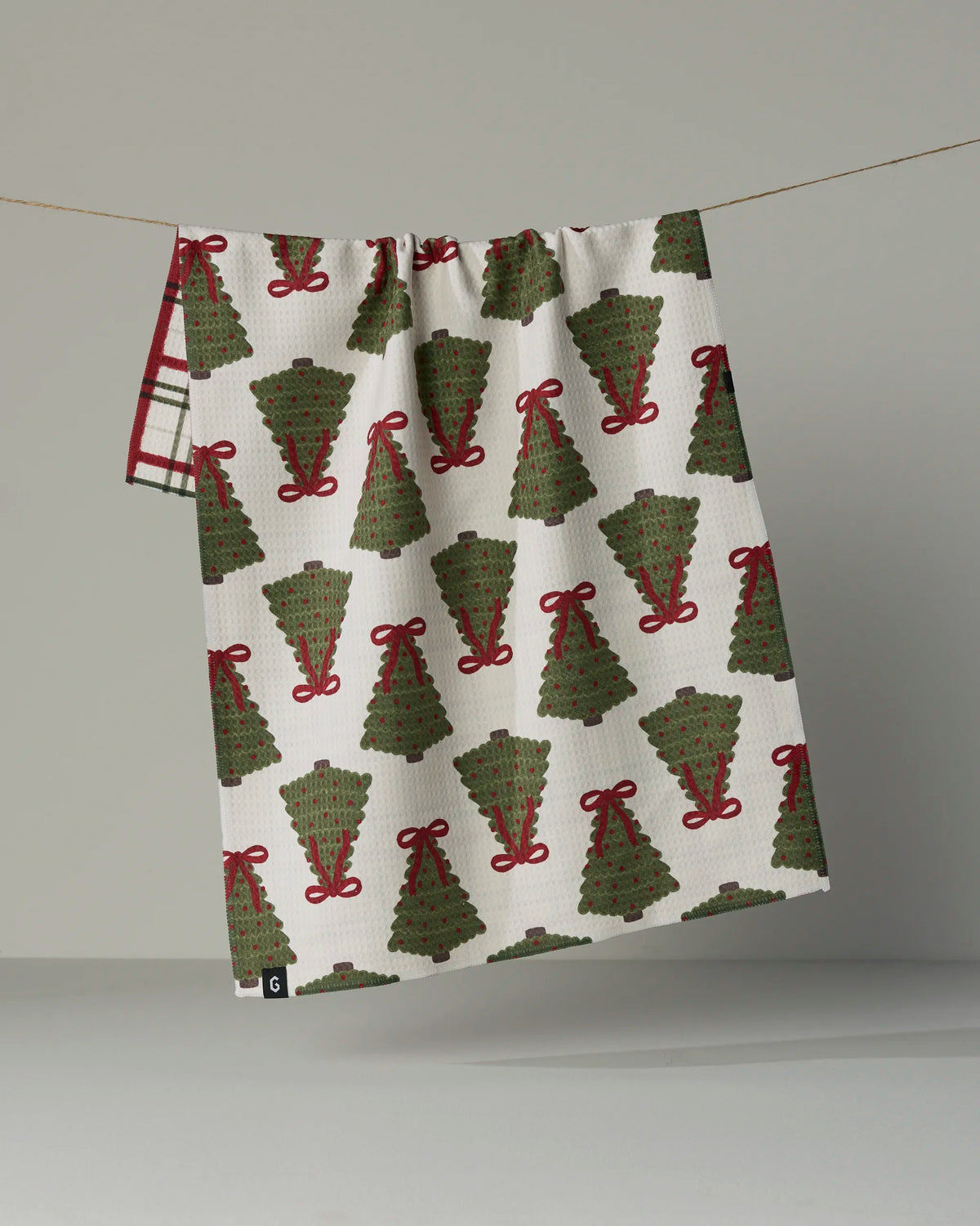A white, quick-drying absorbent towel from Geometry called “Ribbon Pines,” featuring a repeating pattern of green Christmas trees adorned with red bows, hangs on a line against a neutral background.
