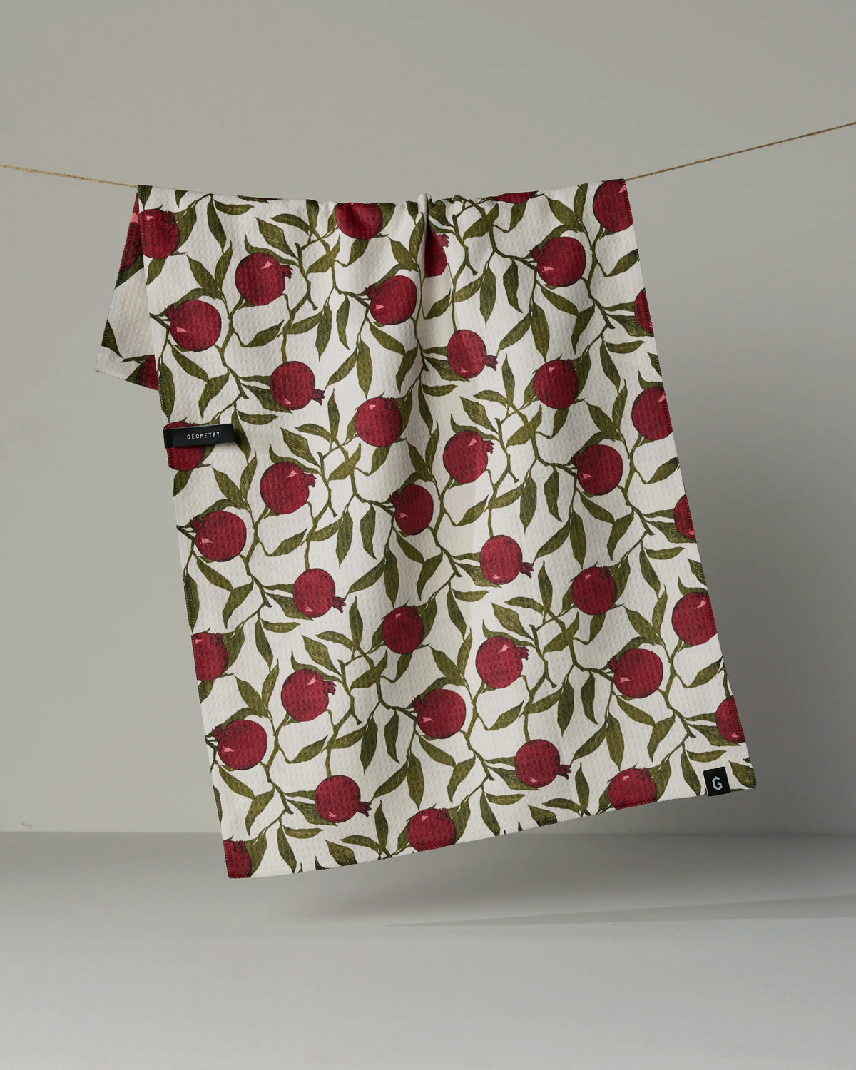 A white, absorbent quick-drying towel from Geometry, called Ruby Rind, featuring a red apple and green leaf pattern hangs on a string against a plain gray background.