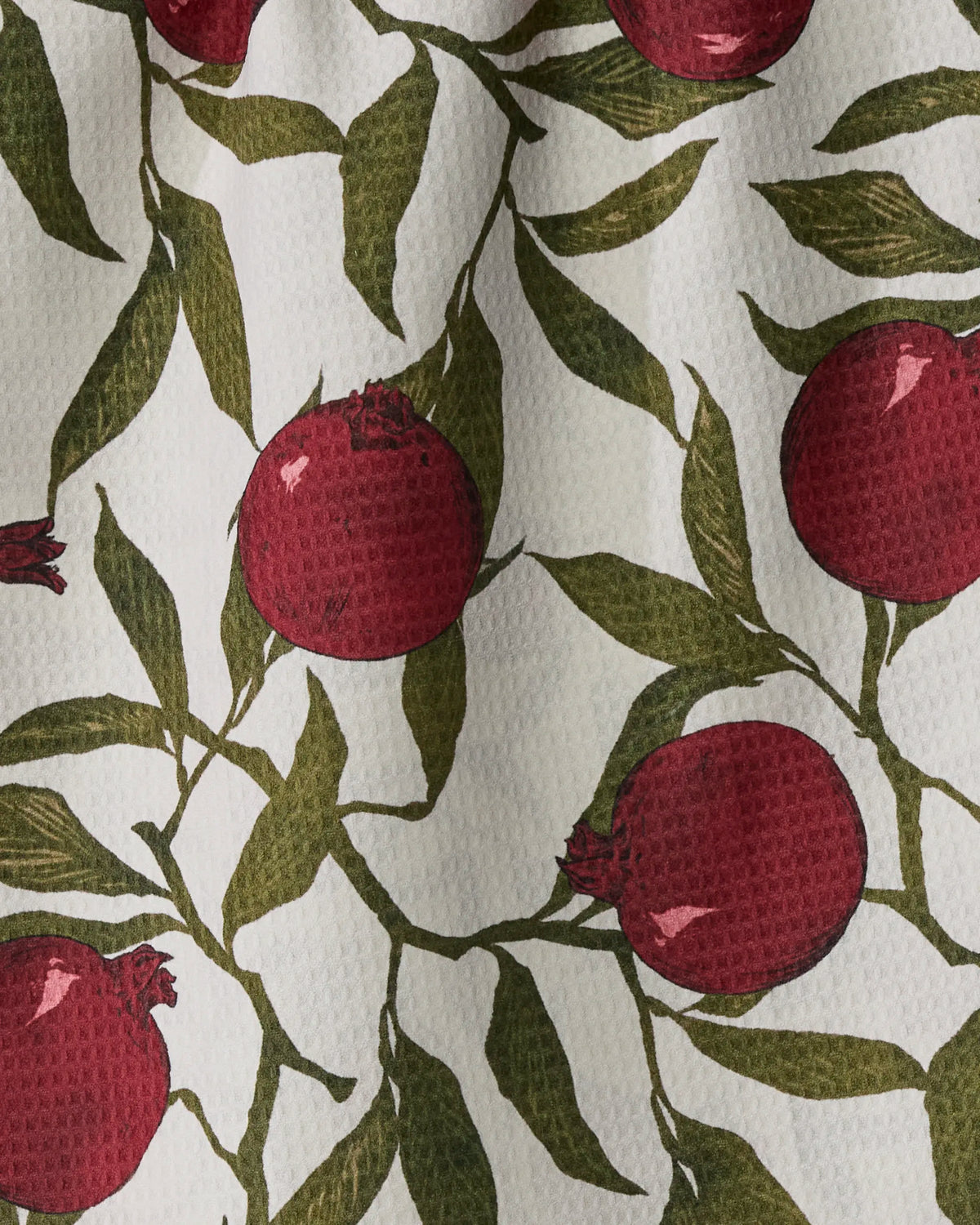 The Ruby Rind towel by Geometry features a textured white surface adorned with a printed pattern of red pomegranates, green leaves, and stems. Its absorbent, quick-drying design combines style and functionality, while the visible dimples enhance both its visual appeal and drying efficiency.