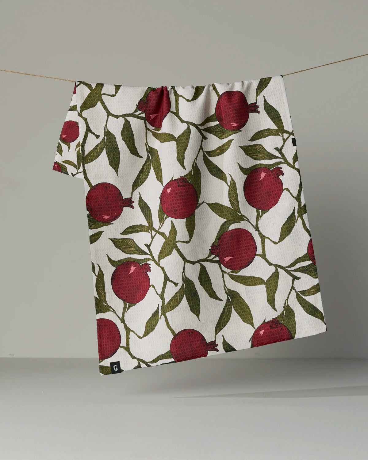 A Ruby Rind tea towel by Geometry, featuring a double-sided white cloth adorned with a pattern of red pomegranates and green leaves, hangs on a string against a plain gray background.