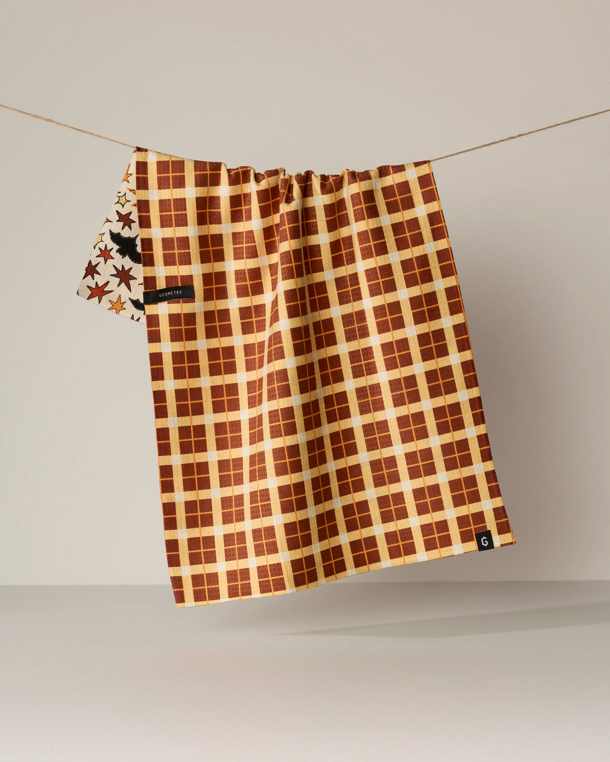 A quick-drying, double-sided tea towel from Geometry, called Sew Batty, hangs on a line. Featuring a brown and yellow plaid pattern with a small star section visible on the left, it’s crafted from recycled materials. The background is plain and neutral.