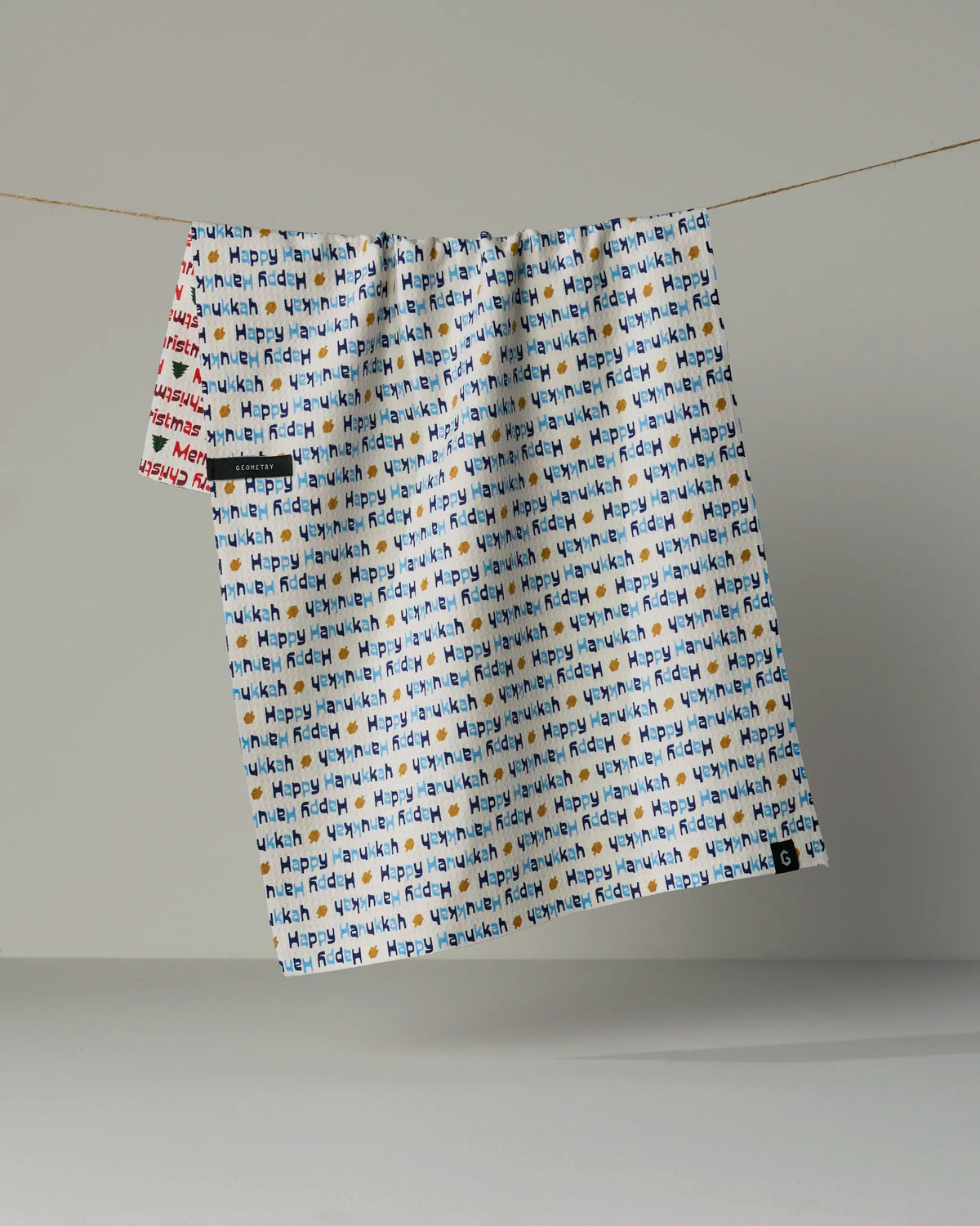 A white, double-sided tea towel from Geometry’s “Shared Joy” collection, featuring a colorful repeating pattern of the words “Shared Joy” in various hues, hangs on a string against a plain gray background. A small section with red text is visible in the upper left corner.