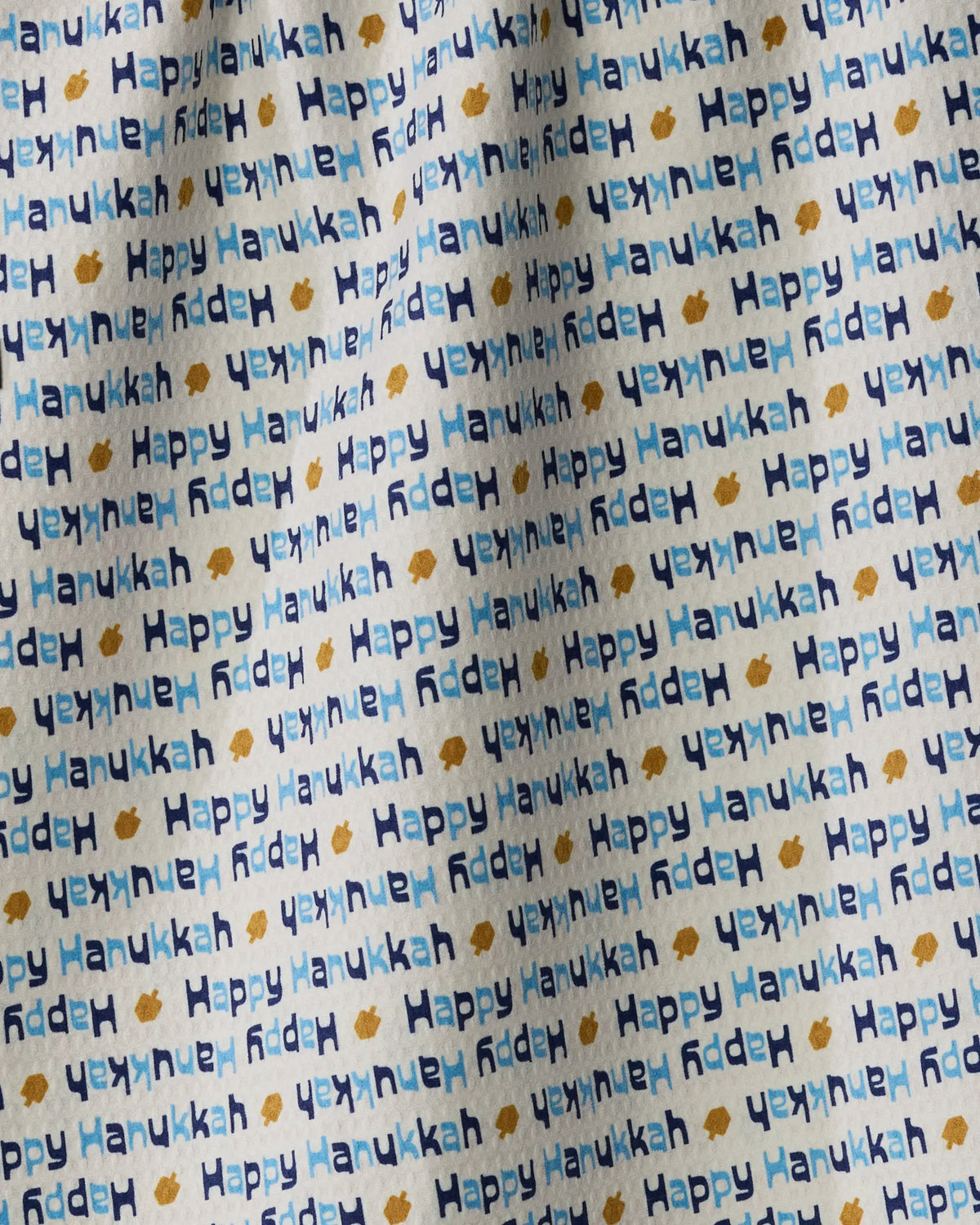 Introducing “Shared Joy” by Geometry: a double-sided tea towel showcasing a wavy patterned fabric adorned with repeating blue and gold “Happy Hanukkah” and “Hidden” text, accented by small gold dreidel icons.