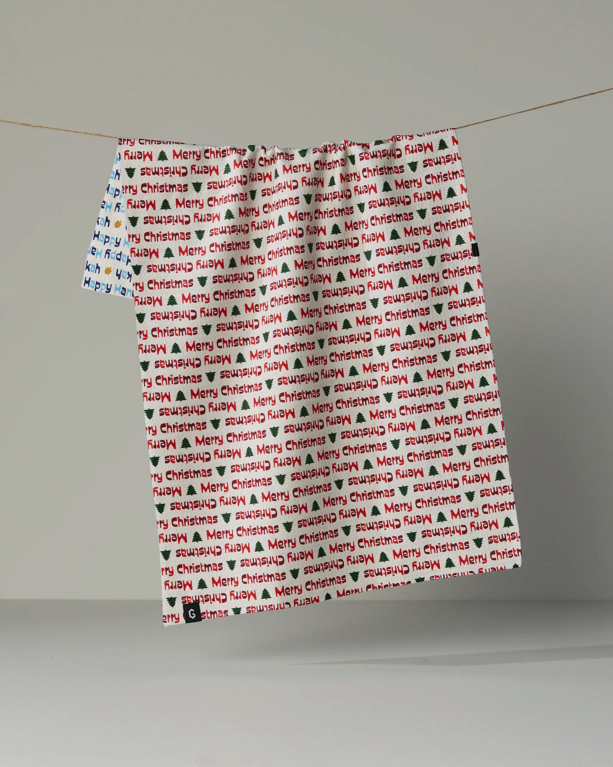 Two sheets from Geometry’s “Shared Joy” collection hang on a string against a plain gray background, each printed with repeating “Merry Christmas” in red, black, and touches of blue. The soft texture is reminiscent of recycled kitchen towels, while subtle lighting casts gentle shadows.