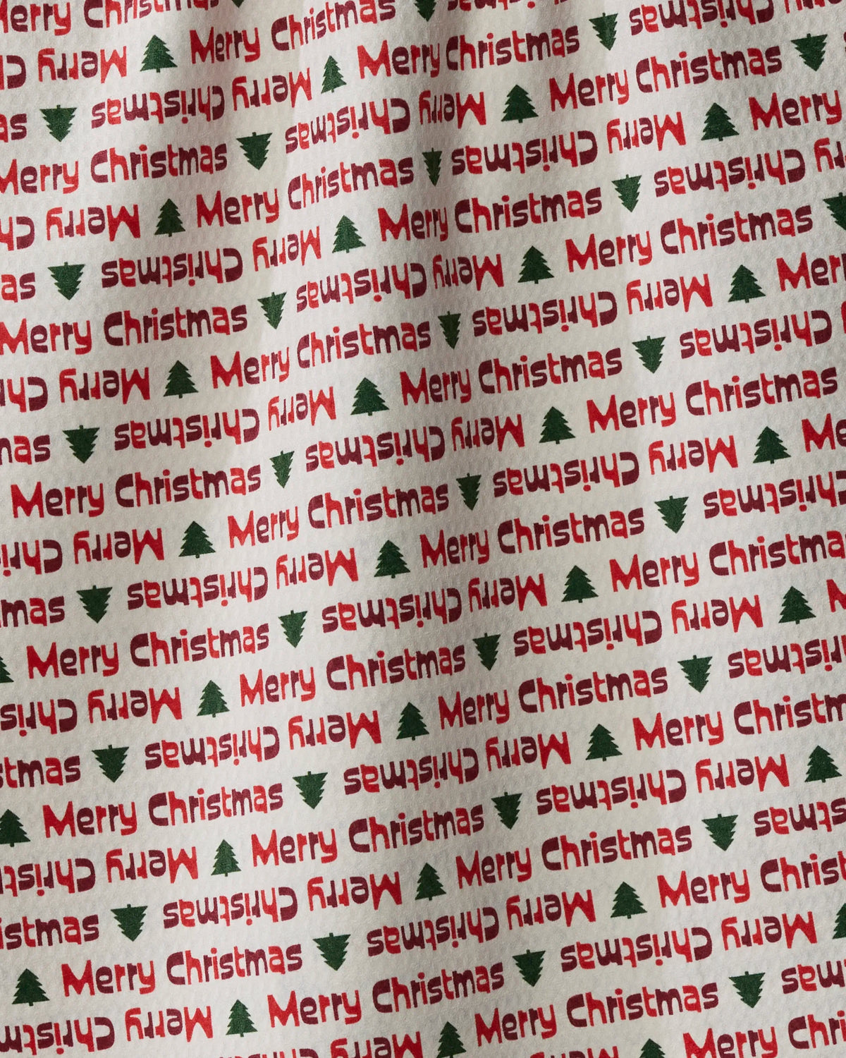 The “Shared Joy” tea towel by Geometry features a festive patterned fabric with “Merry Christmas” in red and small green Christmas trees, displayed in various directions. This double-sided tea towel is perfect for adding holiday cheer to your kitchen while offering both style and versatility.