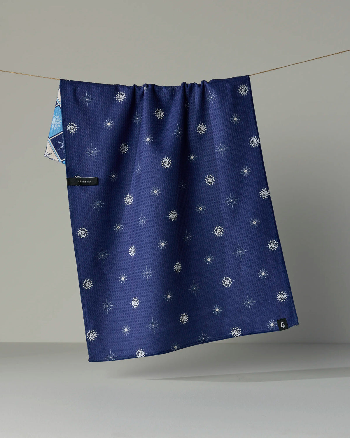 The Geometry Snowflake Chalet tea towel, featuring a navy blue double-sided design with white snowflake patterns, hangs on a clothesline against a neutral background. A small black Geometry label and a folded corner revealing an alternate pattern emphasize its versatile and stylish appeal.