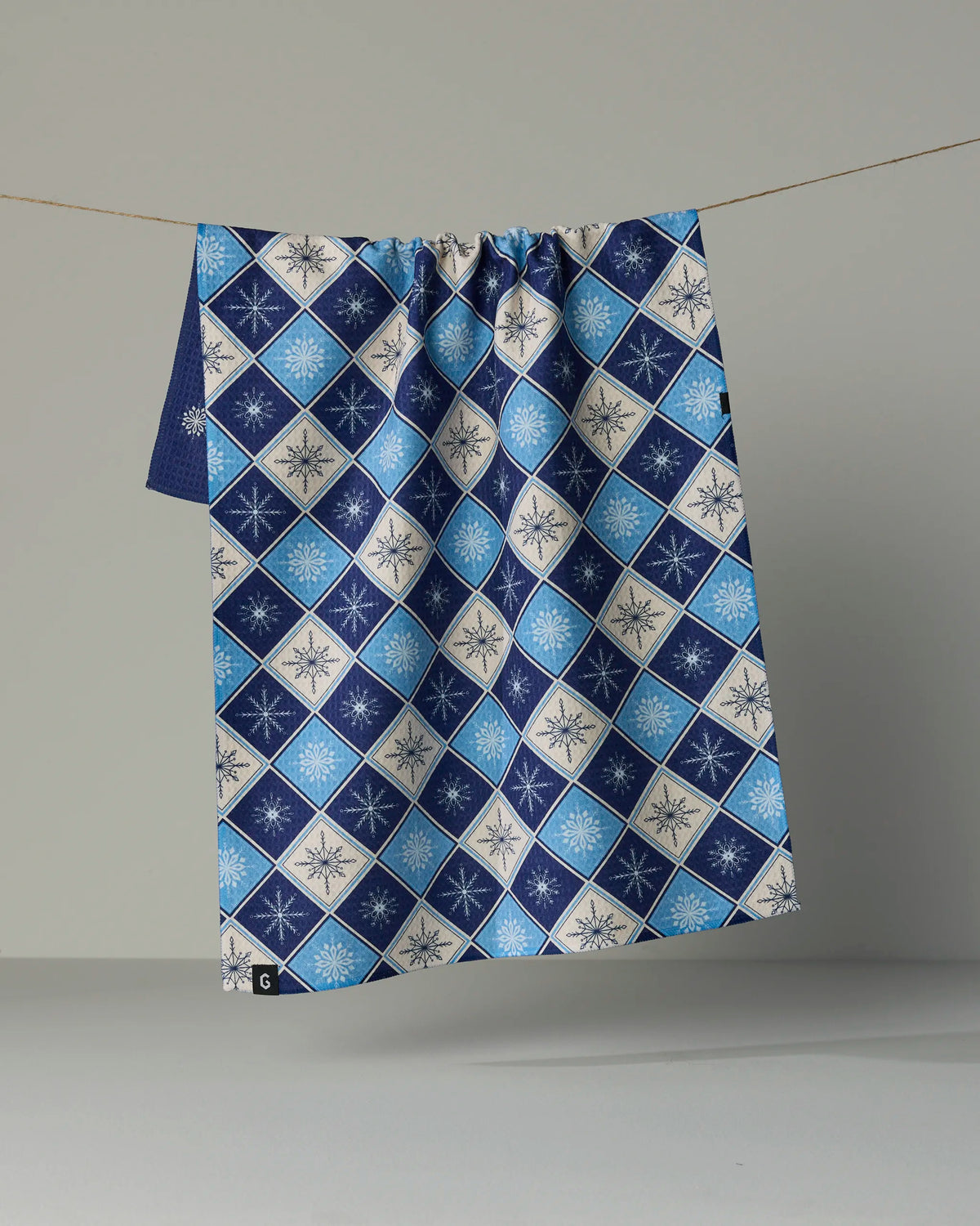 The “Snowflake Chalet” tea towel by Geometry, featuring a blue and white double-sided design with diamond and snowflake patterns, hangs on a clothesline against a plain gray background.