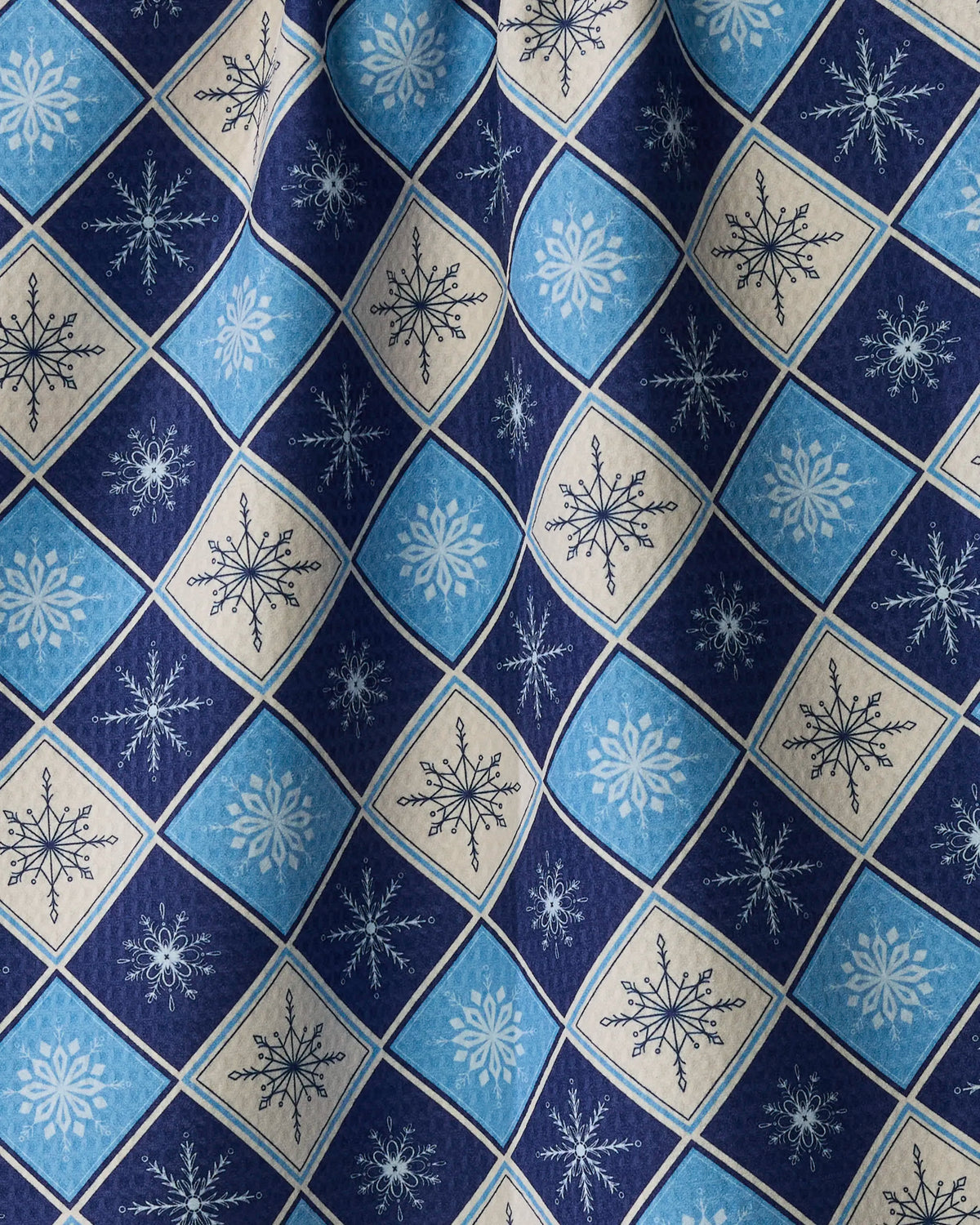 The Snowflake Chalet tea towel by Geometry features blue and beige fabric with a diamond grid pattern adorned with various snowflakes. Crafted from slightly wrinkled, quick-drying material, it creates gentle curved folds across the patterned surface.