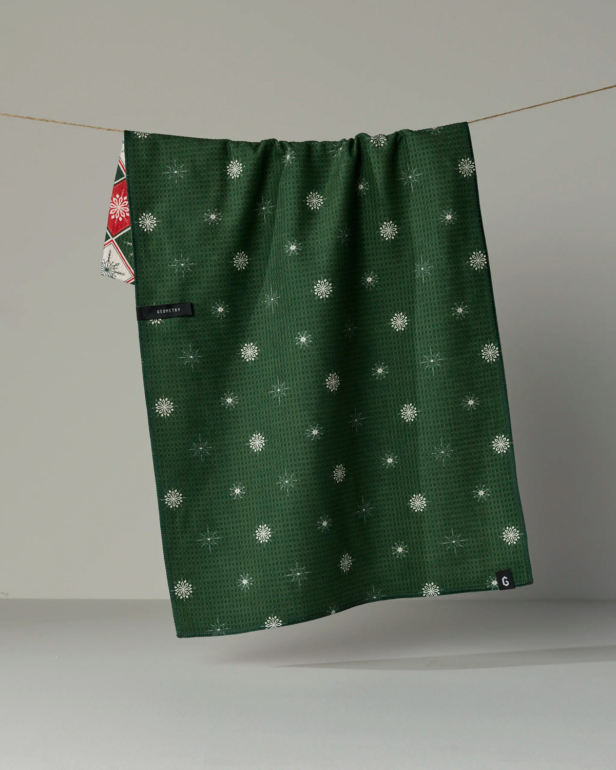 A Geometry “Snowflake Chalet” quick-drying tea towel in green with a white snowflake pattern hangs on a string. A portion of its red and white patterned corner is also visible, set against a plain neutral gray background.