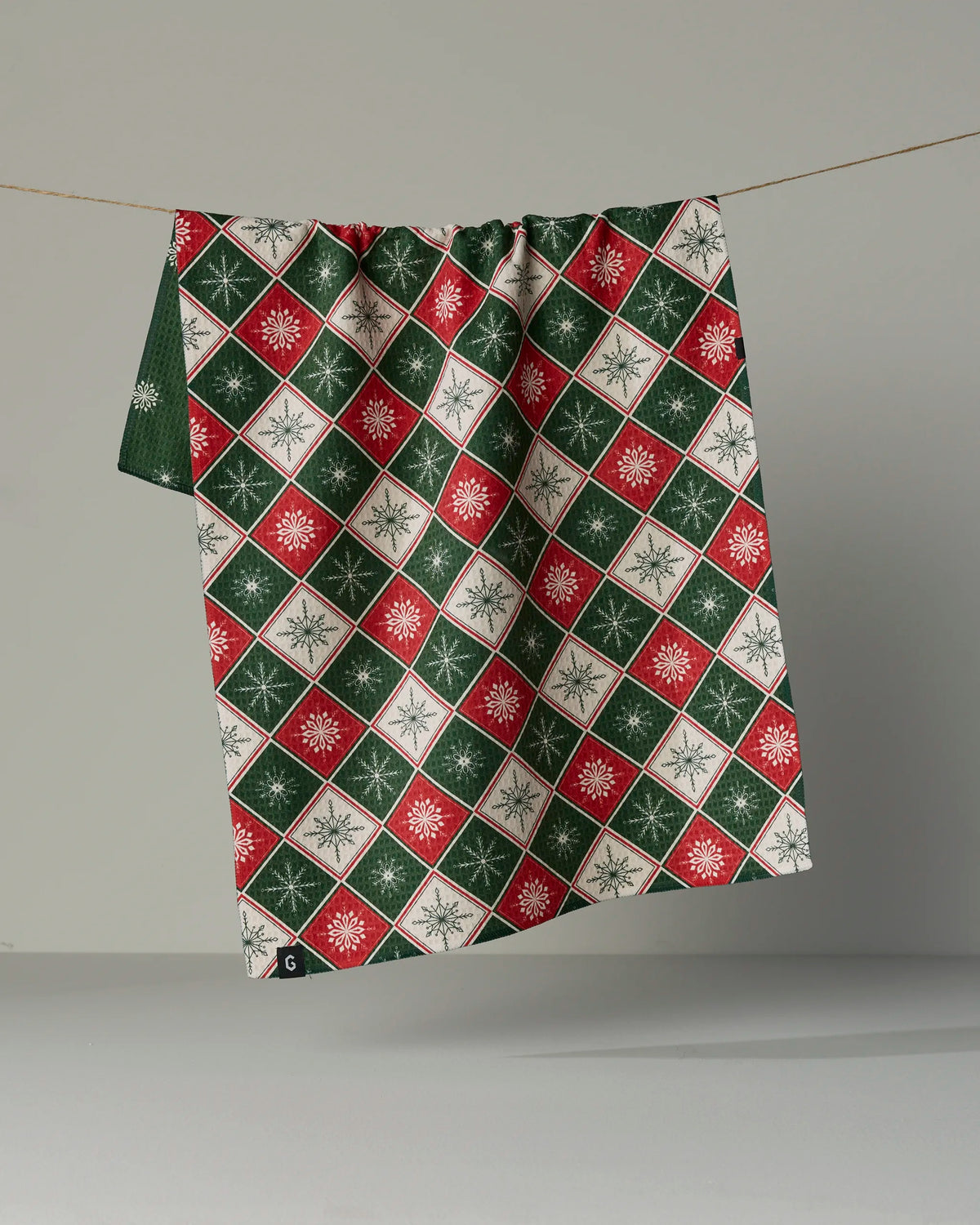 A Geometry Snowflake Chalet recycled kitchen towel, featuring a red, green, and white diamond pattern with white snowflakes, hangs on a string against a plain background.