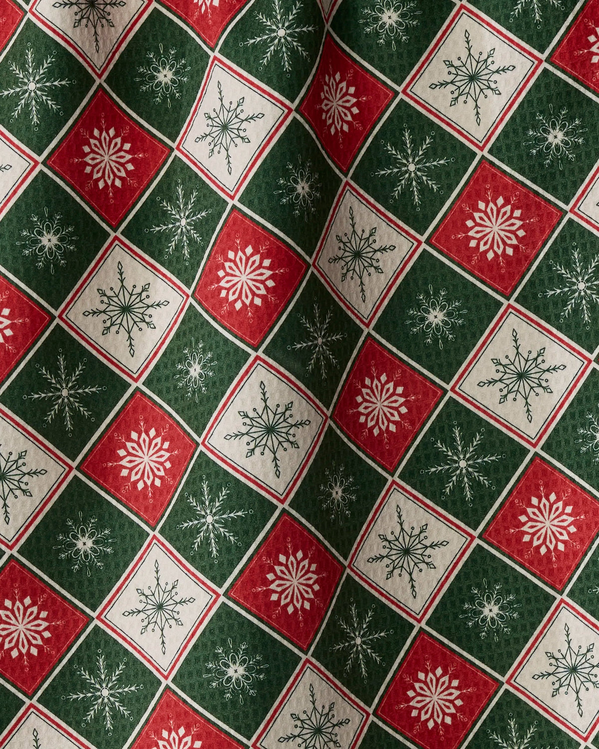 The Geometry Snowflake Chalet tea towel features red, green, and white fabric patterned with alternating squares adorned with snowflake designs. The double-sided festive holiday print, characteristic of the brand’s signature style, appears slightly wrinkled to create subtle shadows and added texture.
