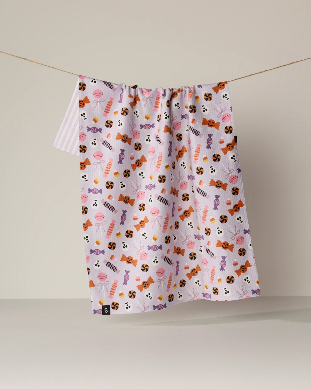 The Geometry “Spooky Sweets” double-sided tea towel, featuring a vibrant pattern of candy, ice cream, and sweets in orange, purple, and black, hangs on a clothesline against a plain beige background.
