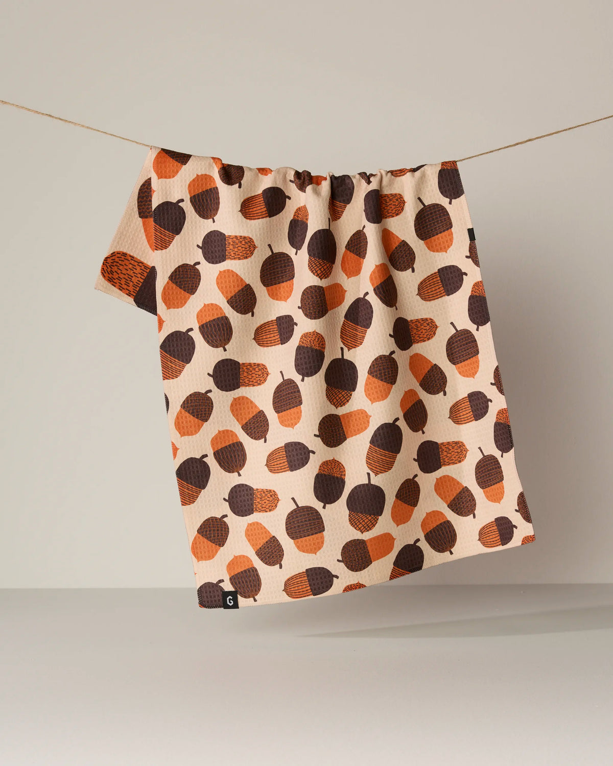 The Squirrel Bounty tea towel by Geometry, featuring a brown and orange acorn pattern on beige, hangs on a string against a plain background.