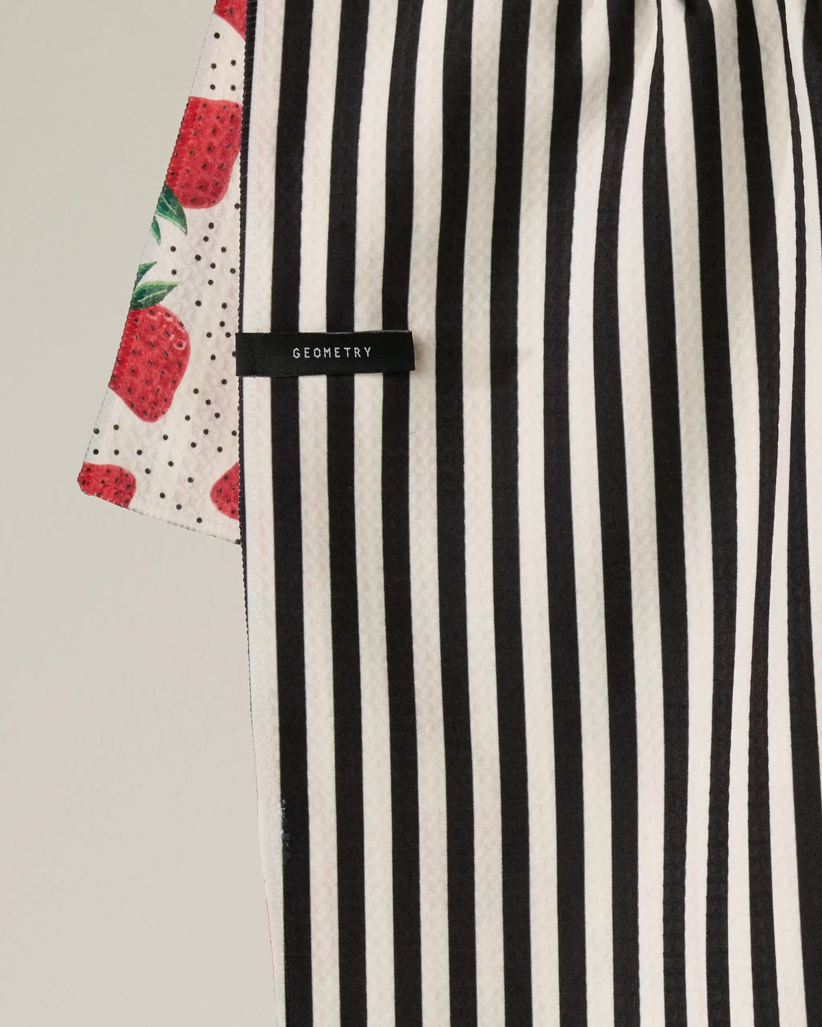 A recycled kitchen towel with vertical black and white stripes and a visible Geometry label overlaps the “Strawberry Lane” towel by Geometry, which features a pattern of red strawberries and small black dots on a cream background.