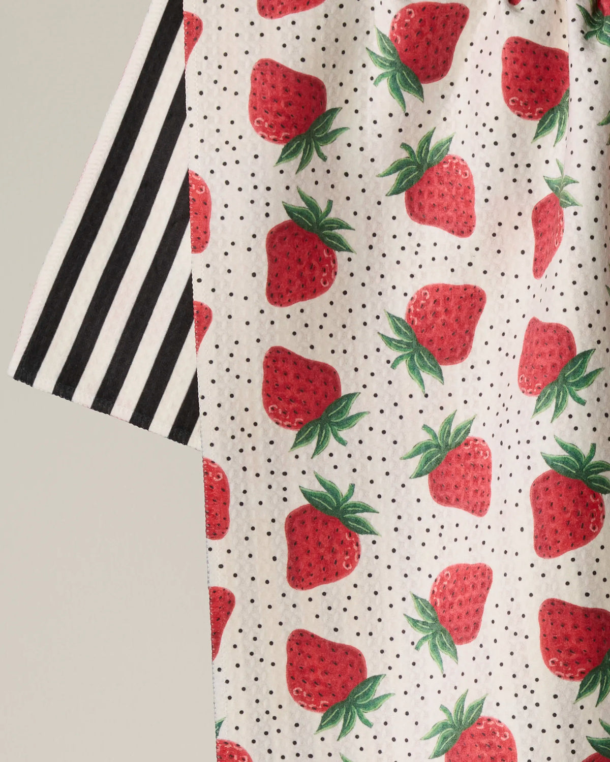 The Geometry Strawberry Lane tea towel features a quick-drying fabric adorned with a playful pattern of red strawberries and small black dots on a crisp white background, accented by a strip of vertical black-and-white stripes along one edge.