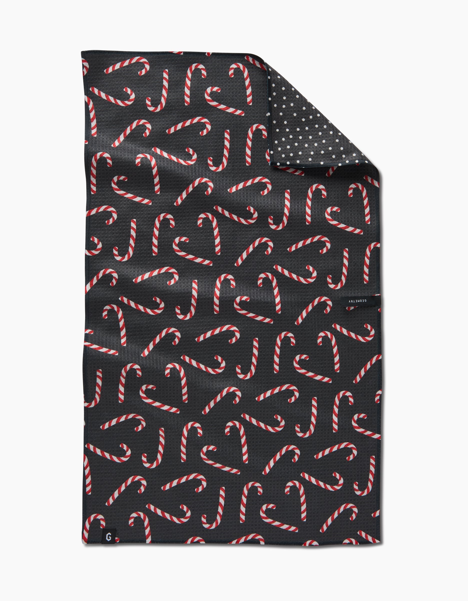 The Geometry Sweet Canes Double-Sided Tea Towel features a black cloth with red and white candy canes, plus a folded corner revealing a gray underside with white polka dots. Made from recycled materials.