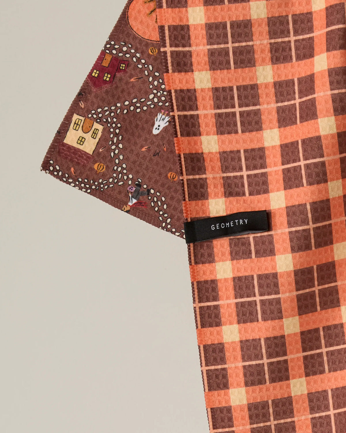 Two overlapping cloths: one is the Geometry “Treat Trail” tea towel, featuring a Halloween village pattern with houses, a ghost, and a pumpkin; the other is an orange and brown plaid quick-drying tea towel with a black Geometry tag visible.