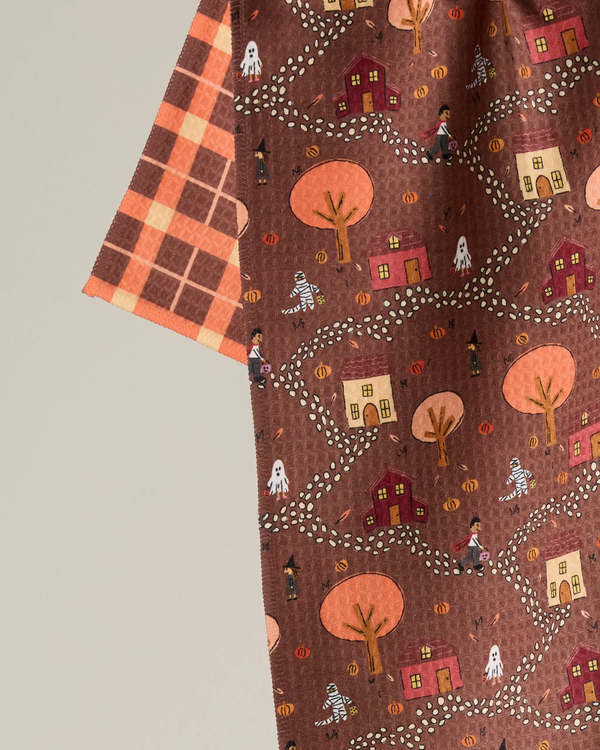 The “Treat Trail” tea towel by Geometry features a patterned brown fabric adorned with autumn trees, houses, ghosts, mummies, pumpkins, and costumed children—a charming design for this double-sided tea towel. A contrasting orange plaid edge on the left side adds a playful touch to this quick-drying towel, making it perfect for festive kitchens.