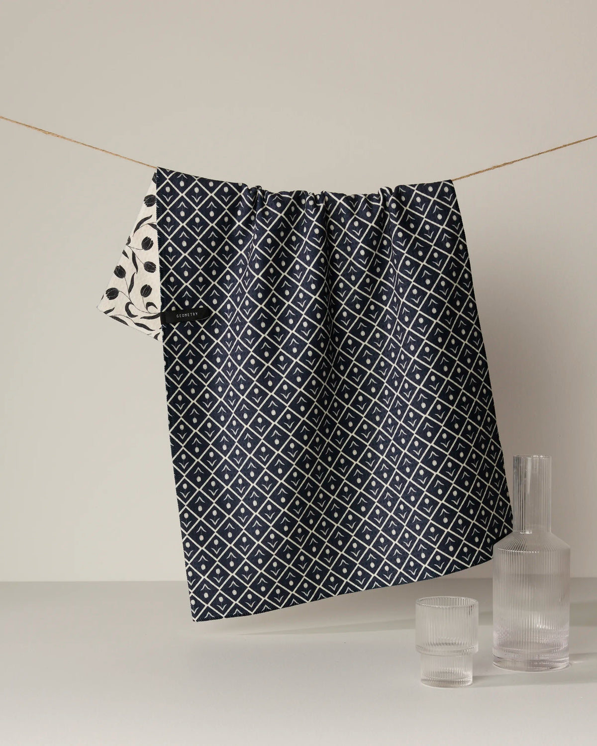 A Tulip Noir double-sided tea towel from Geometry, featuring a navy blue and white geometric pattern, hangs on a string with a glimpse of white and black fabric beside it. Below, three clear ribbed glass vases and cups rest on a white surface.