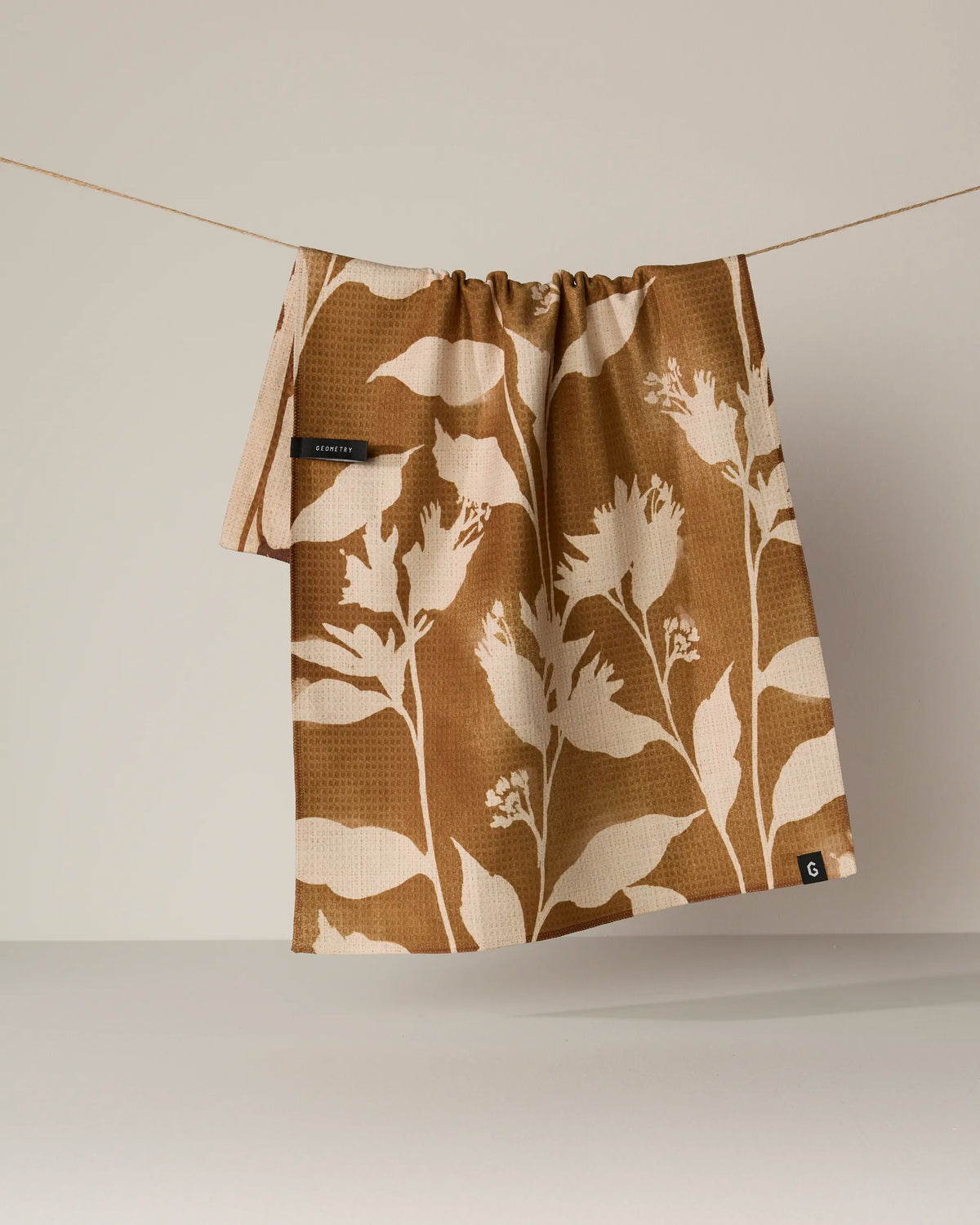 The Geometry Willowmere tea towel, featuring a brown and beige double-sided floral pattern, hangs on a clothesline against a neutral background.