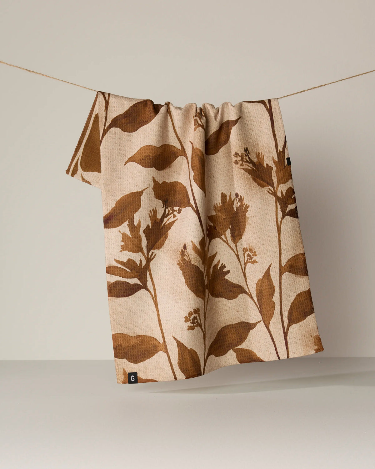 A Willowmere recycled kitchen towel from Geometry, featuring a brown botanical leaf and flower pattern on beige, hangs on a thin string against a neutral background.
