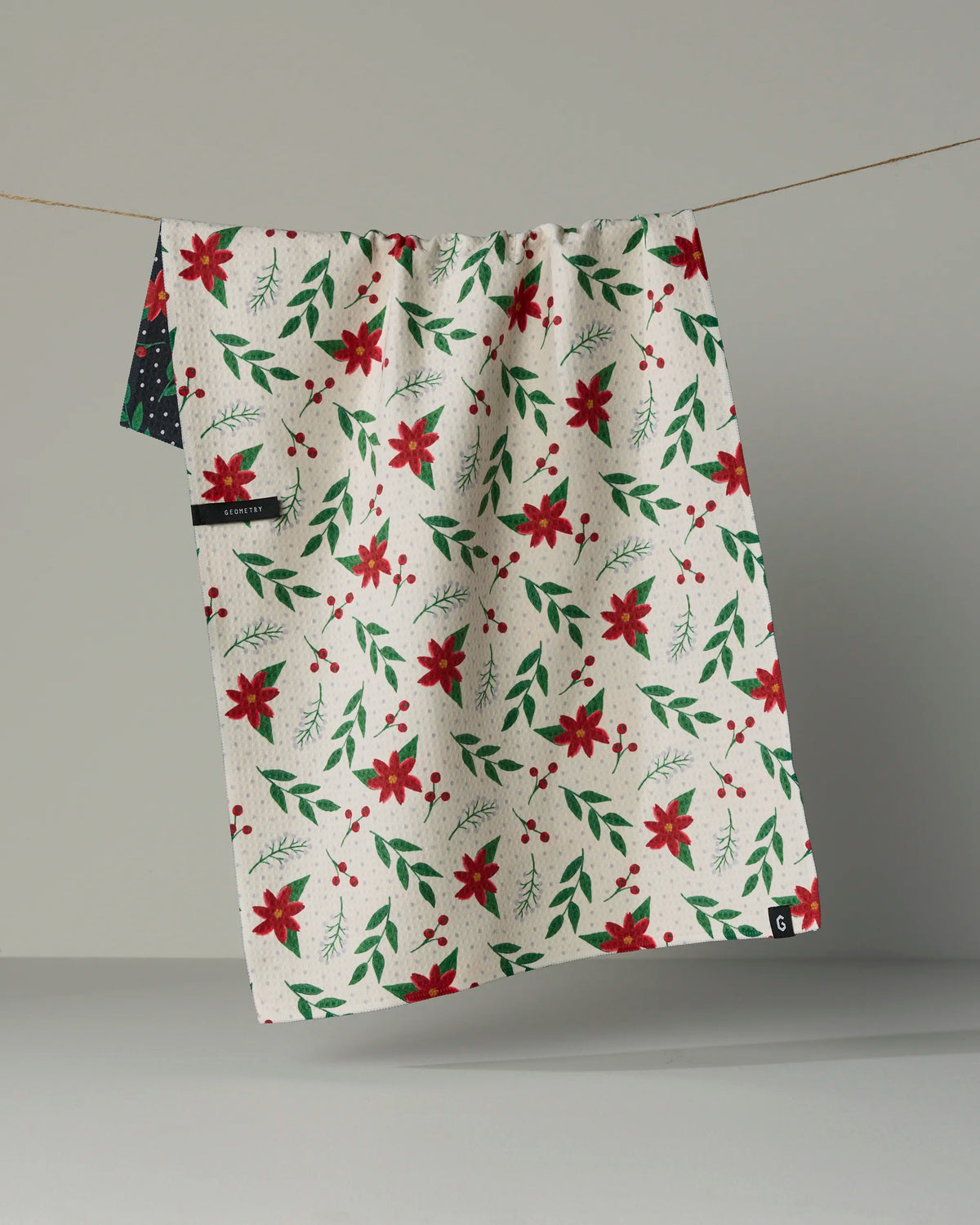 The Winter Bloom tea towel from Geometry features a festive double-sided design with red poinsettias, green leaves, and small red berries, elegantly draped over a line against a neutral background. Made from recycled materials, this quick-drying and eco-friendly cloth adds stylish holiday charm to your home.