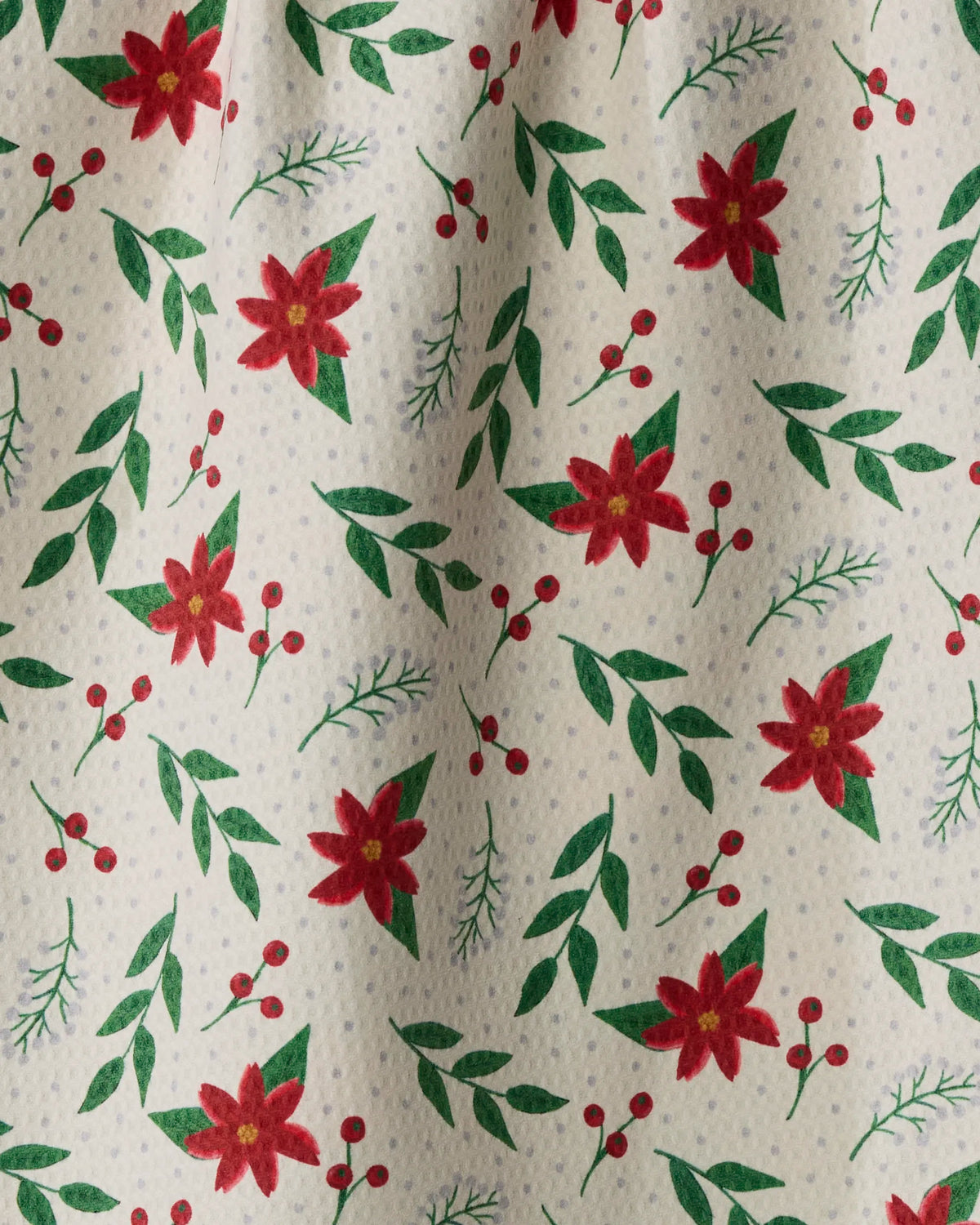 Winter Bloom by Geometry is a festive tea towel crafted from recycled materials, featuring a vibrant pattern of red poinsettia flowers, green leaves, sprigs, and red berries on a light background. This slightly wrinkled double-sided design merges sustainability with seasonal charm.
