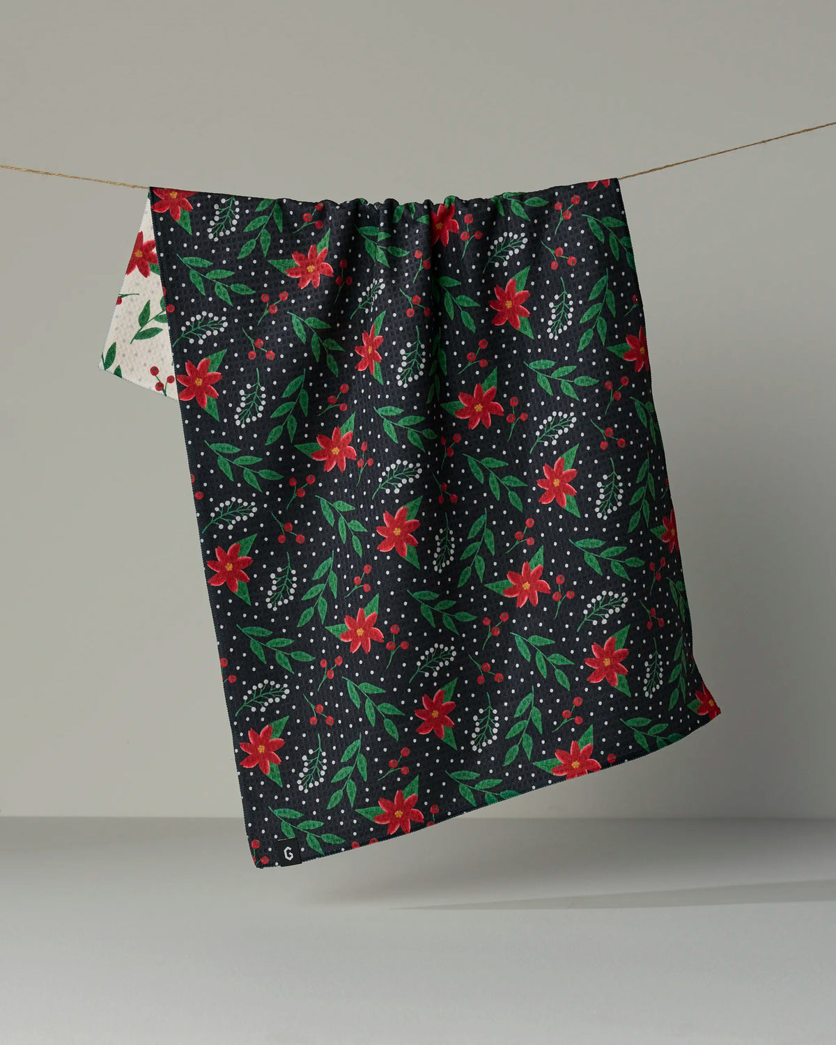 A quick-drying, double-sided Geometry “Winter Bloom” tea towel made from recycled materials hangs on a string, its dark background decorated with a pattern of red flowers, green leaves, and white dots, while the reverse side reveals a lighter floral design.