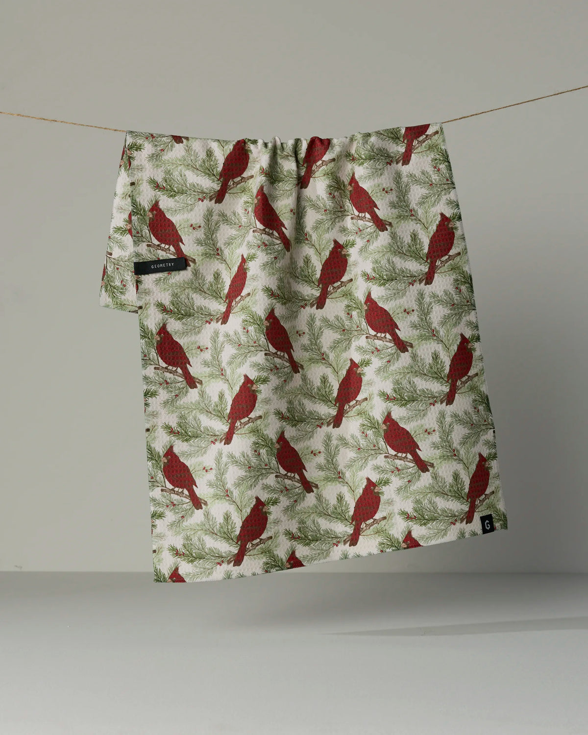 The Winter Crimson tea towel by Geometry, featuring a double-sided design of red cardinals perched on green pine branches, hangs on a line against a plain, light background.