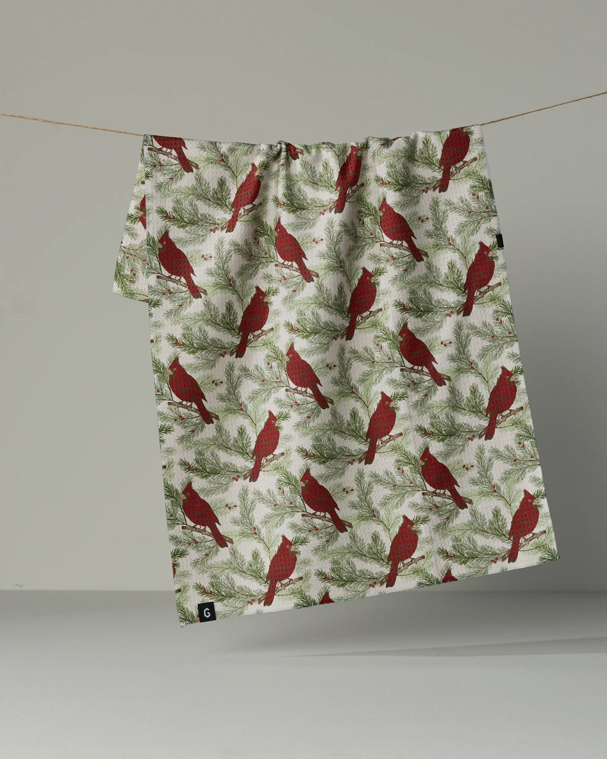 The Winter Crimson tea towel by Geometry, featuring a red cardinal and green pine branch design on both sides, hangs on a clothesline against a plain, light-colored background.