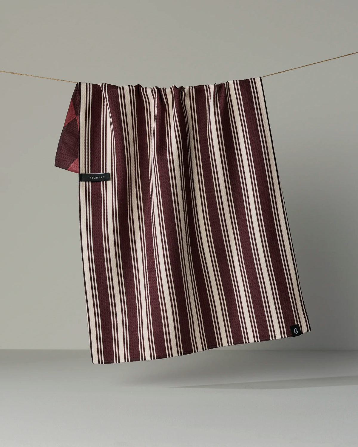 A maroon and white striped double-sided tea towel from Geometry, called “Winter Gems,” hangs from a thin line against a plain gray background, featuring quick-drying properties for everyday convenience.
