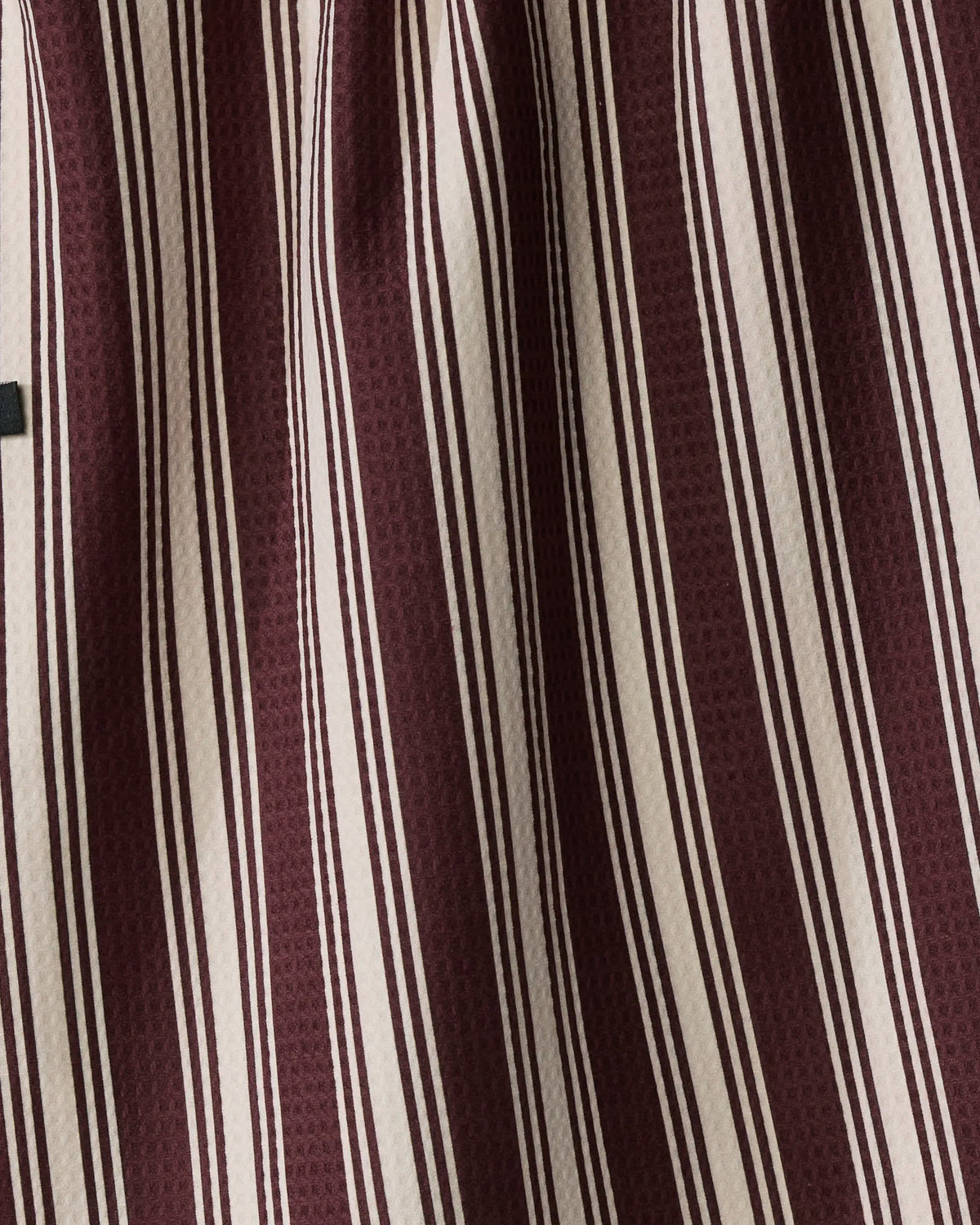 A close-up of Geometry’s “Winter Gems” fabric reveals vertical maroon and beige stripes, expertly crafted from recycled materials. The textured material, with its subtle folds and shadows, makes it ideal for a double-sided tea towel.