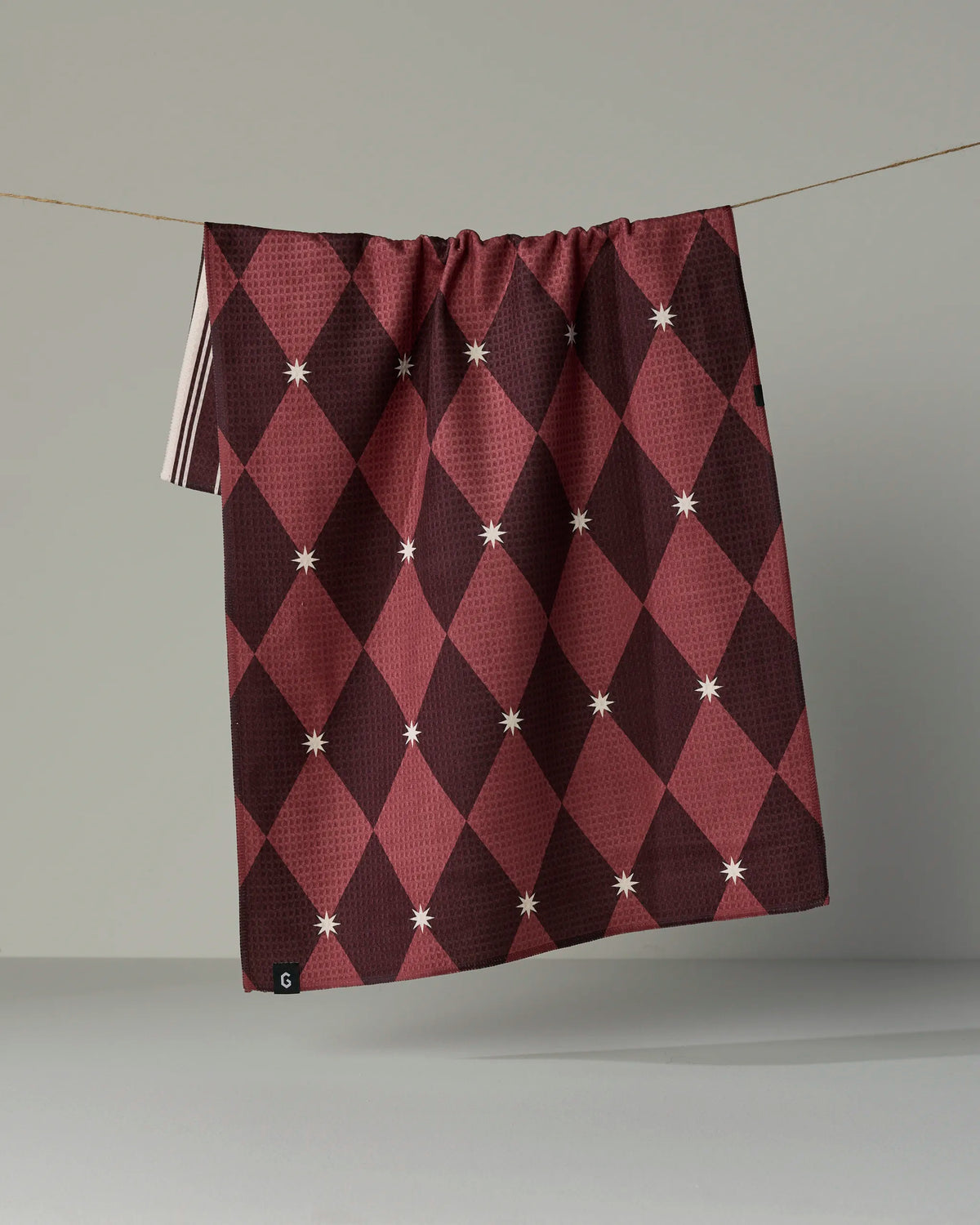 Geometry’s “Winter Gems” is a maroon and dark red diamond-patterned cloth, adorned with small white star accents and crafted from recycled materials, displayed hanging on a clothesline against a plain gray background.