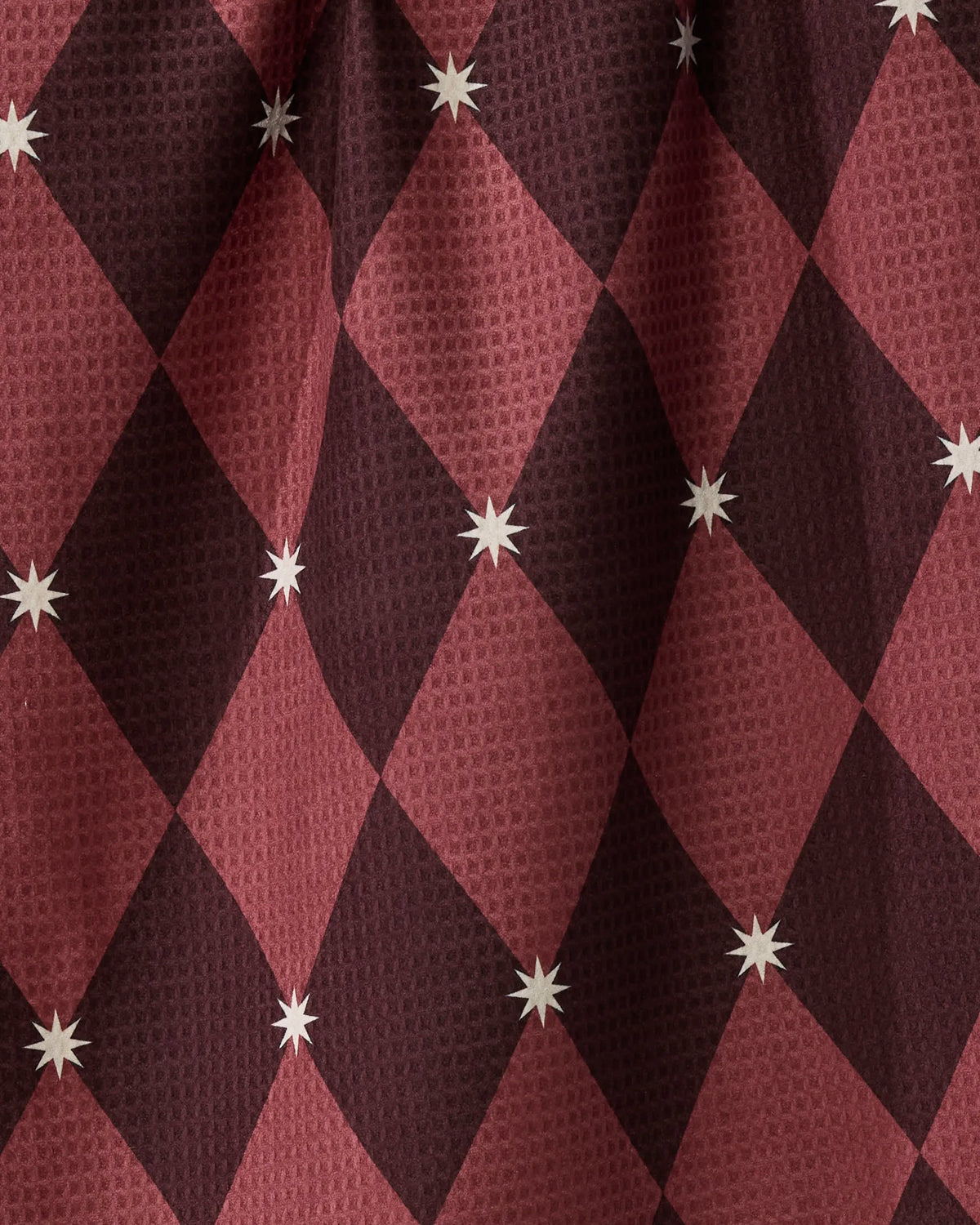 Winter Gems by Geometry features a maroon and burgundy diamond-patterned fabric adorned with small white eight-pointed stars at select intersections. With its subtly textured, slightly wrinkled surface, this double-sided tea towel is crafted from recycled materials—seamlessly blending sustainable design with everyday practicality.