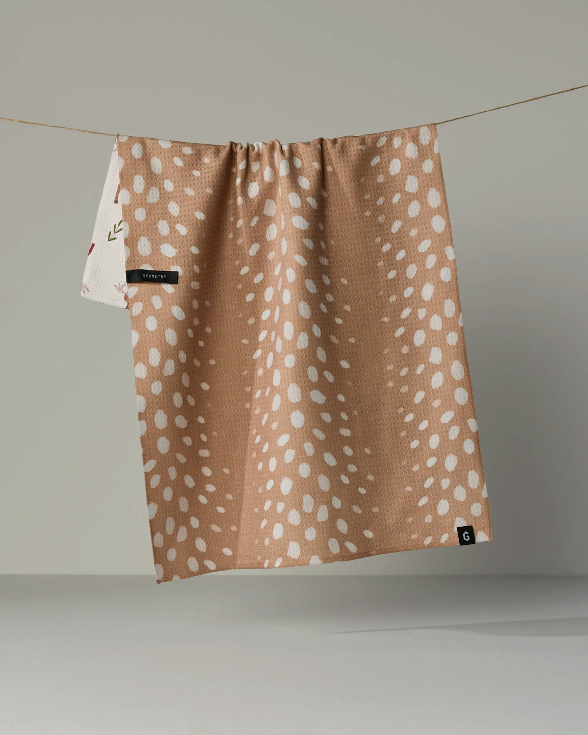 A tan, quick-drying Geometry tea towel called “Winter Reindeer,” adorned with white spotted patterns and a small black tag on the bottom corner, hangs on a clothesline with part of another white towel visible in the background.