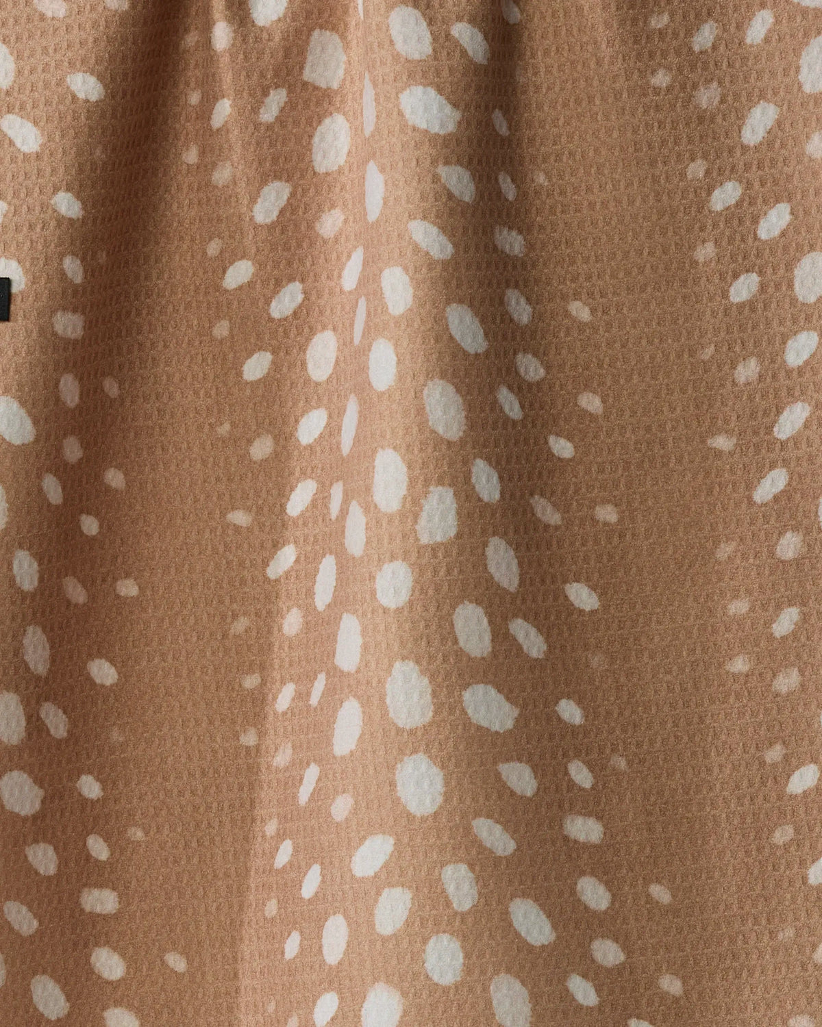 Close-up of the Geometry Winter Reindeer tea towel shows its tan fabric adorned with a white spotted pattern reminiscent of reindeer fur. The textured surface features tiny, raised bumps that emphasize the tea towel’s absorbency and quick-drying qualities. A small black rectangular detail is partially visible along the left edge.