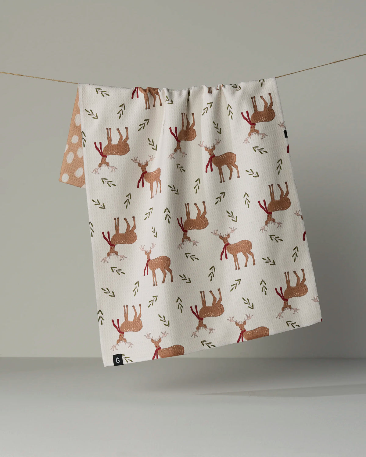 The Geometry “Winter Reindeer” tea towel, featuring a quick-drying beige fabric adorned with red and brown reindeers and small green arrows, hangs on a thin string against a plain light gray background.